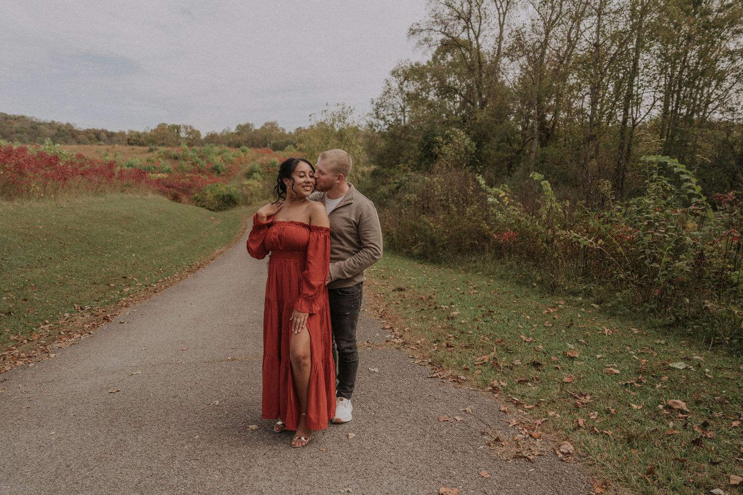 A couple shares a tender moment on a paved path surrounded by autumn foliage and greenery.