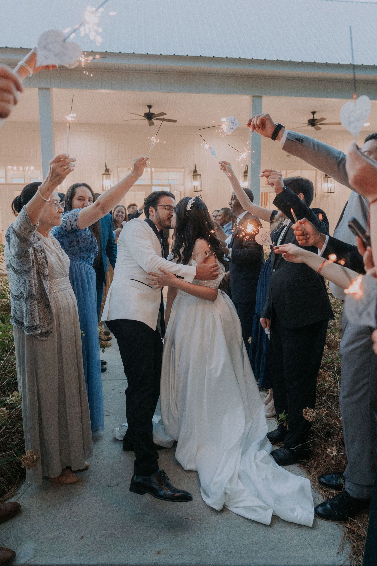 Bride and groom share a kiss during a romantic sparkler send-off surrounded by cheering wedding guests.