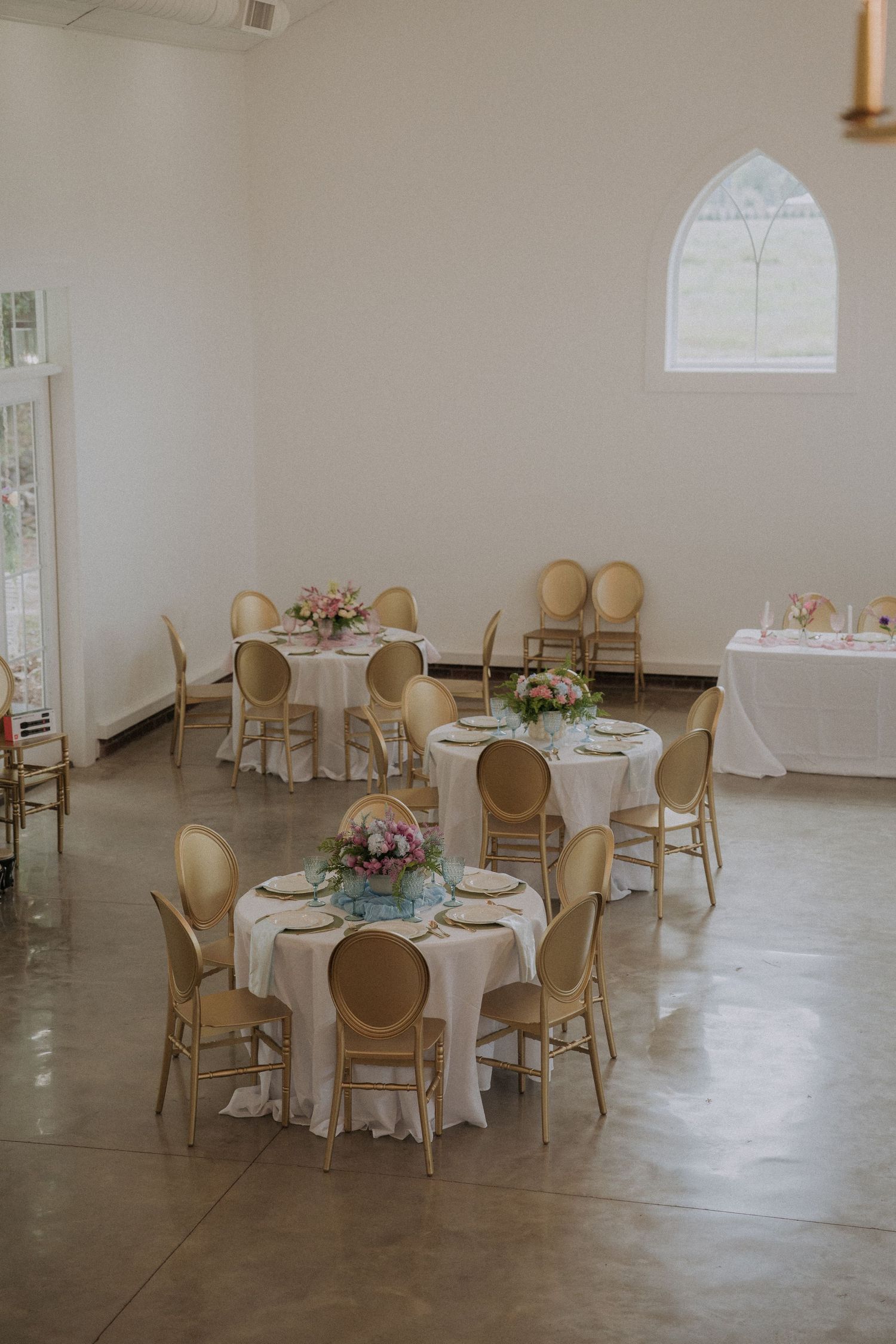 Elegant wedding reception hall with round tables, white linens, gold chairs, and floral centerpieces in a bright white venue.