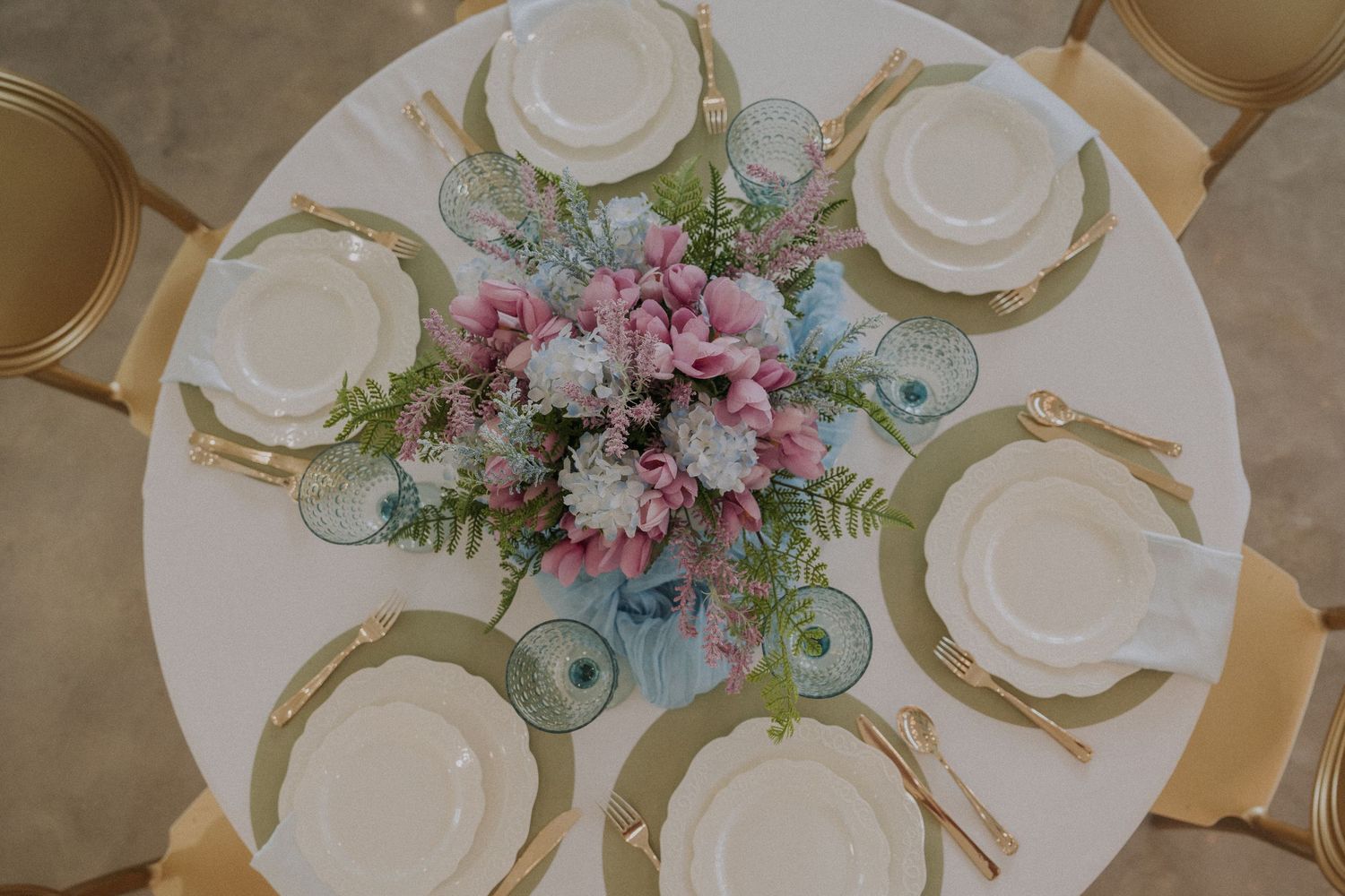 Elegant round wedding table with pink floral centerpiece, white plates, gold cutlery, and green fern accents, viewed from above.