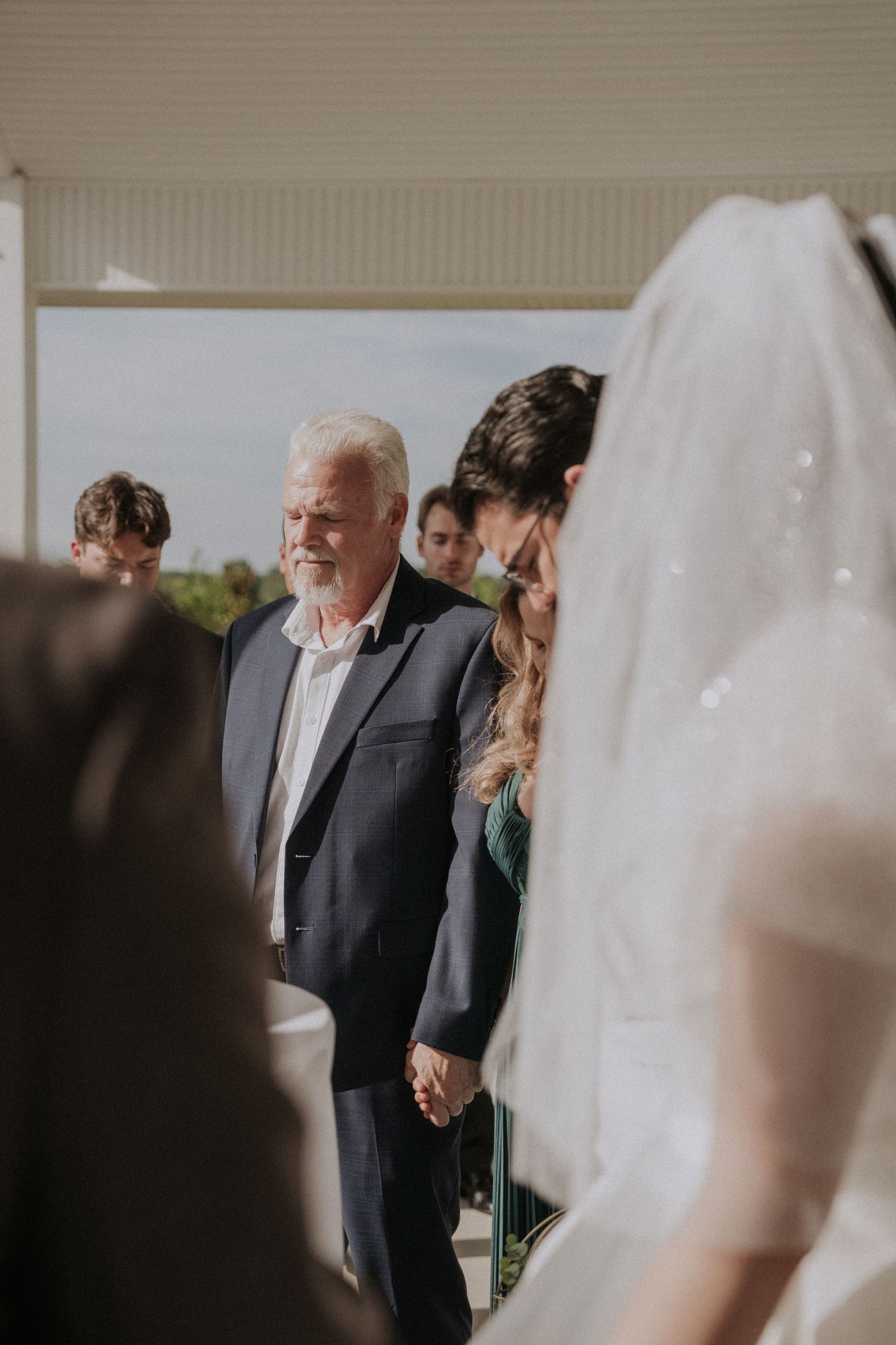 Elderly man in navy suit holds bride's hand during outdoor wedding ceremony procession.
