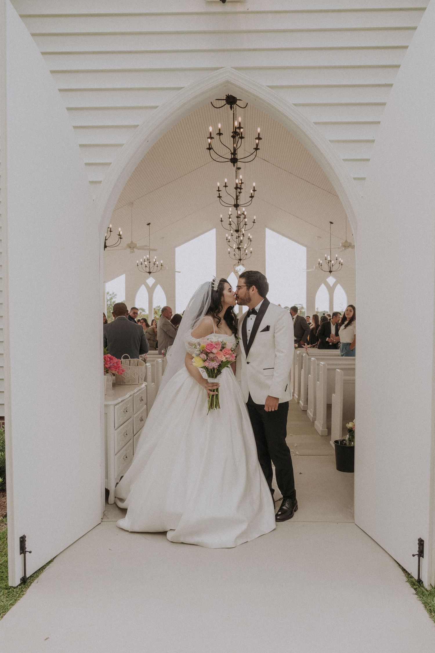 Bride and groom share a kiss under a white Gothic arch in an elegant chapel, bride holding a colorful bouquet.