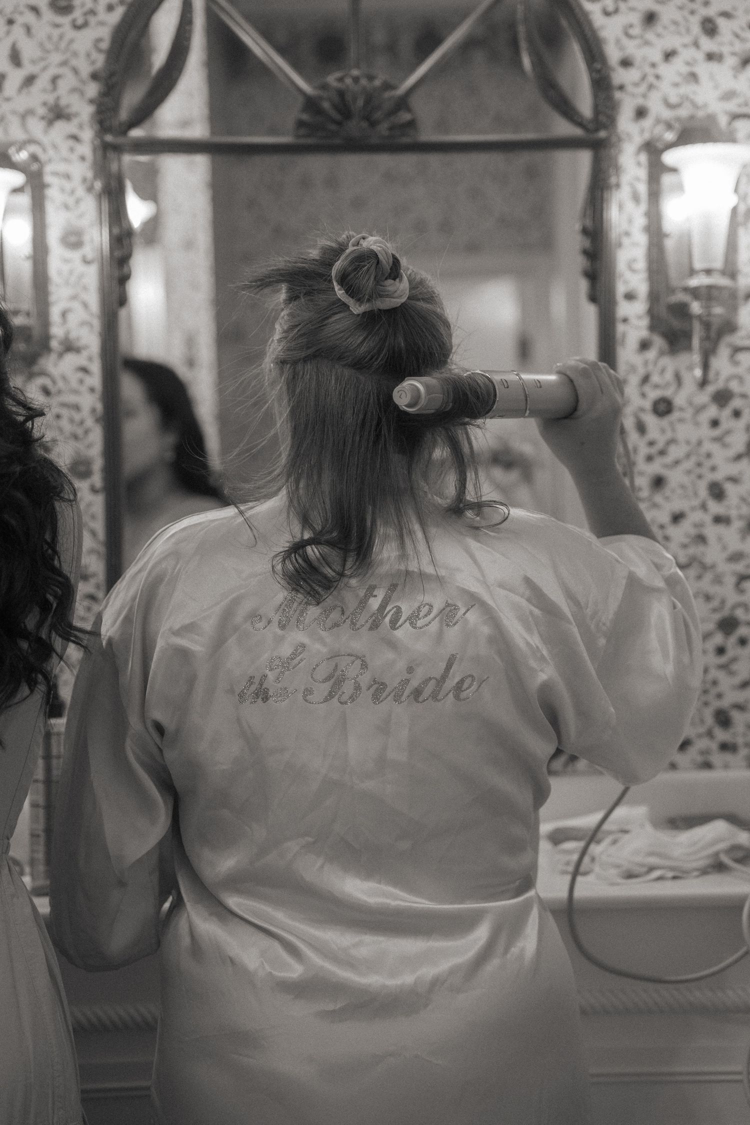 Mother of the bride in white robe styling hair with curling iron before wedding ceremony, black and white photo.