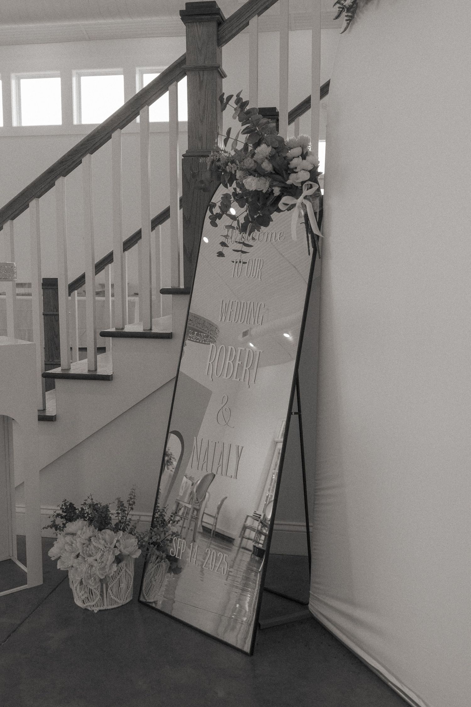 Black and white photo of a tall mirror wedding welcome sign for Robert and Nataly, adorned with flowers, beside a staircase.