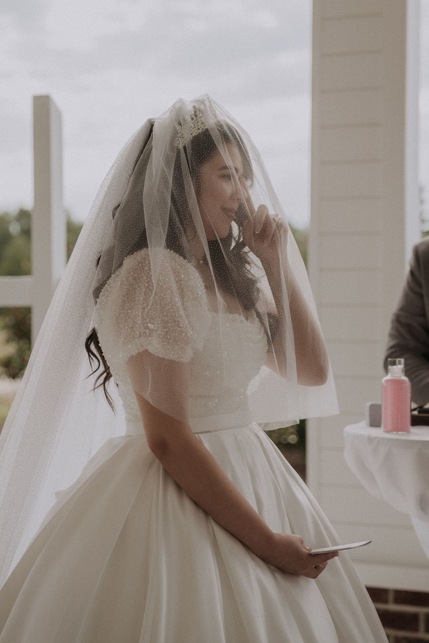 A bride in a ballgown and veil wipes a tear during her outdoor wedding ceremony.