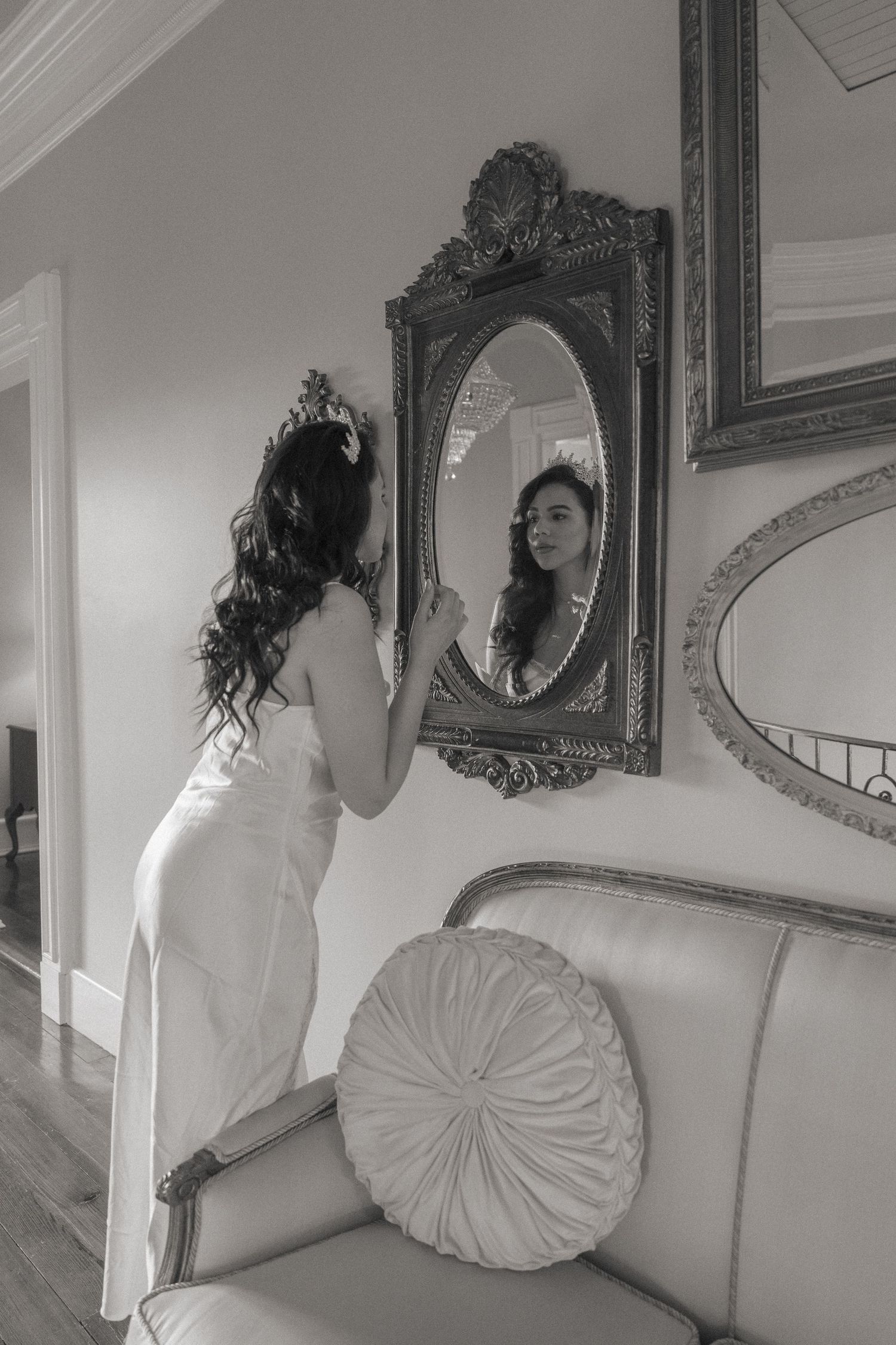 Black and white photo of a bride in a white gown admiring herself in an ornate gold mirror.