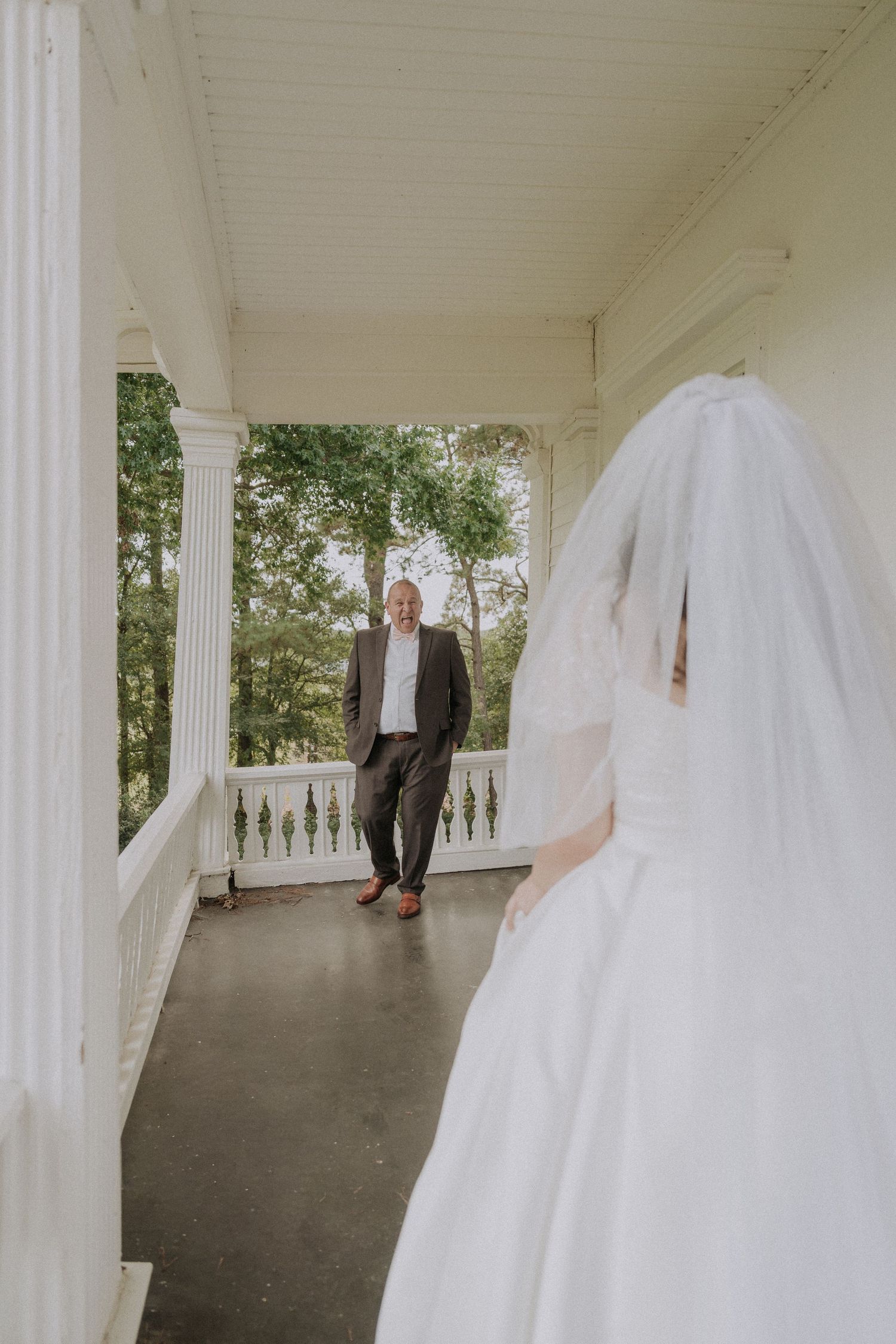 Bride in white gown and veil faces smiling groom on classic white columned porch.