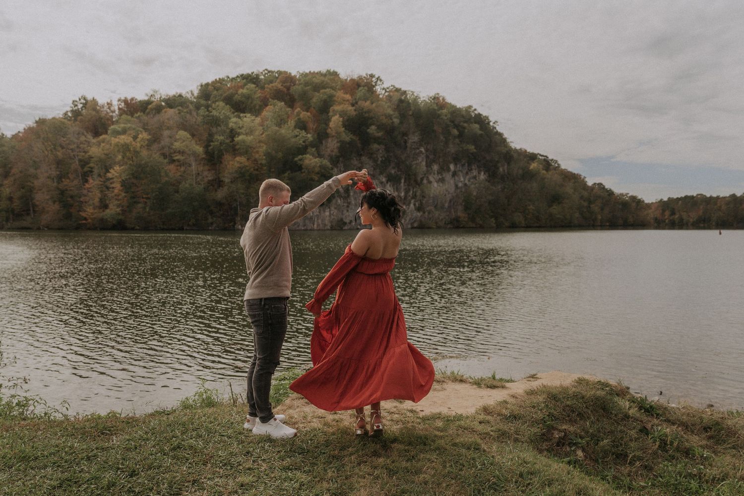 Couple dancing by a serene lake with autumn trees, woman in flowing red dress, romantic outdoor engagement photo.