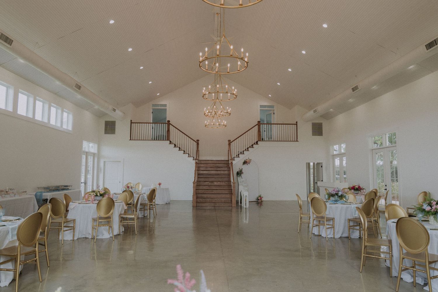 Elegant wedding reception hall with chandeliers, gold chairs, white tables, and grand staircase in a bright venue.