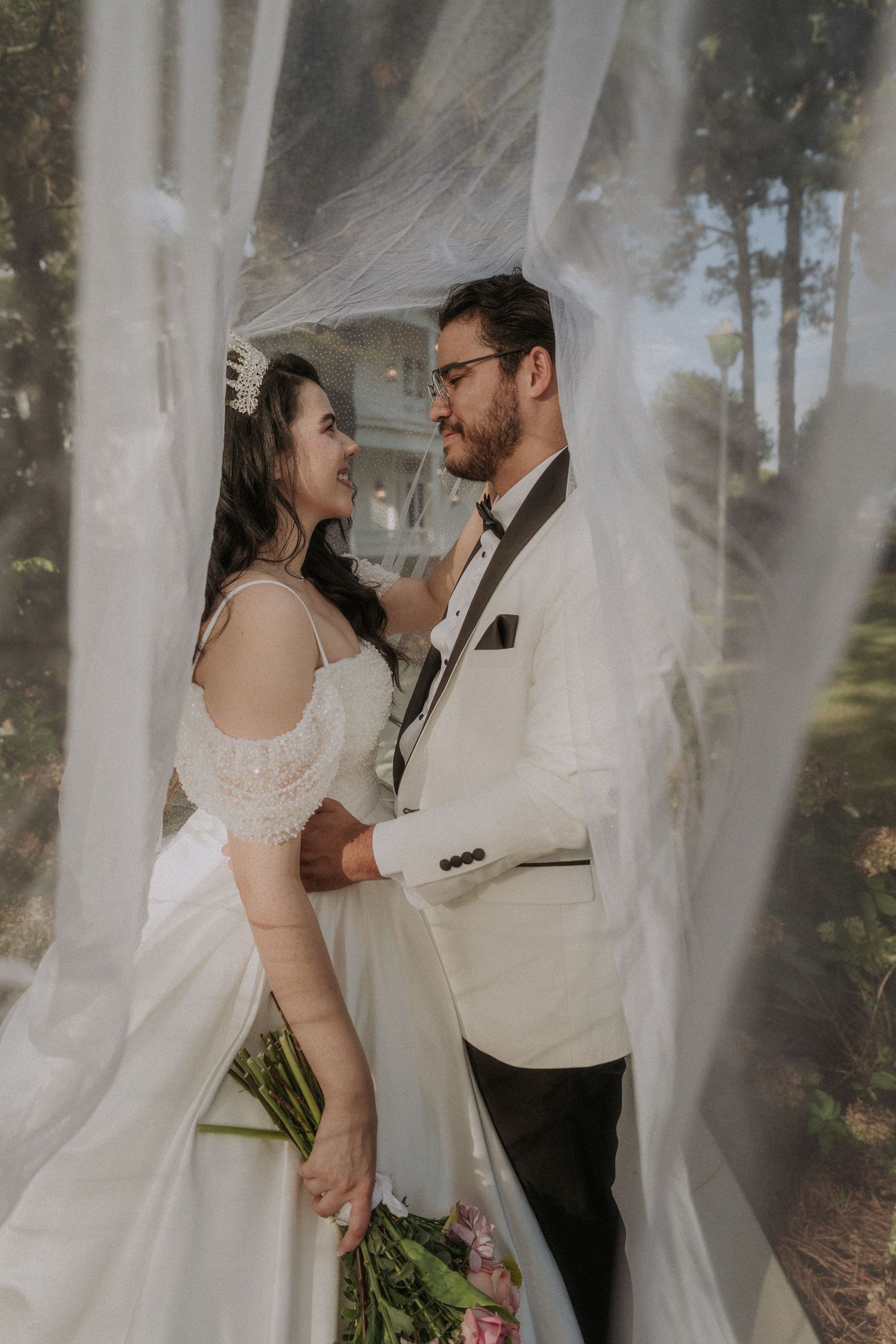 Bride and groom share a romantic moment framed by flowing white curtains at an outdoor wedding venue.