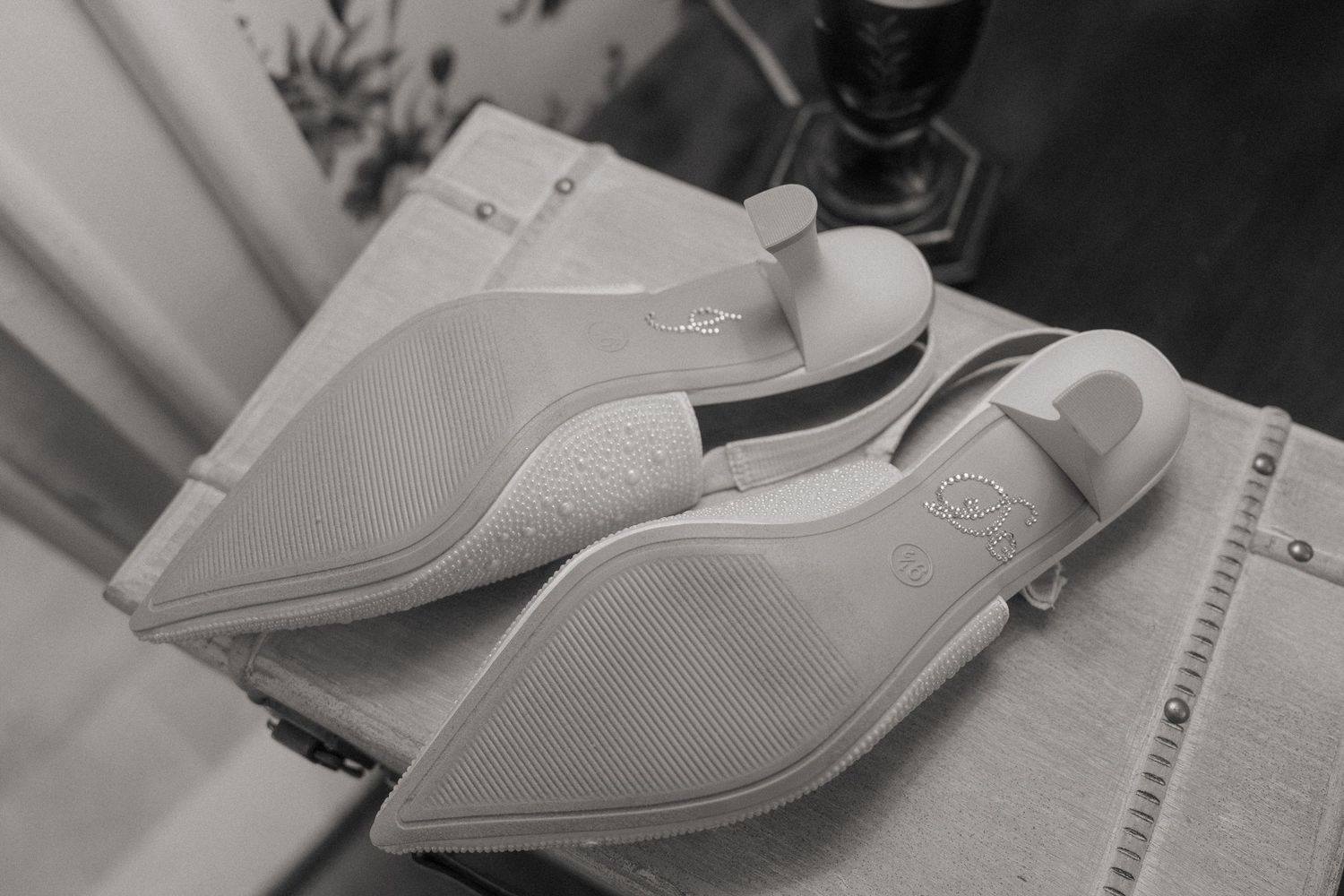 Black and white photo of white bridal heels displayed sole-up on a soft surface, showing elegant embossed details.