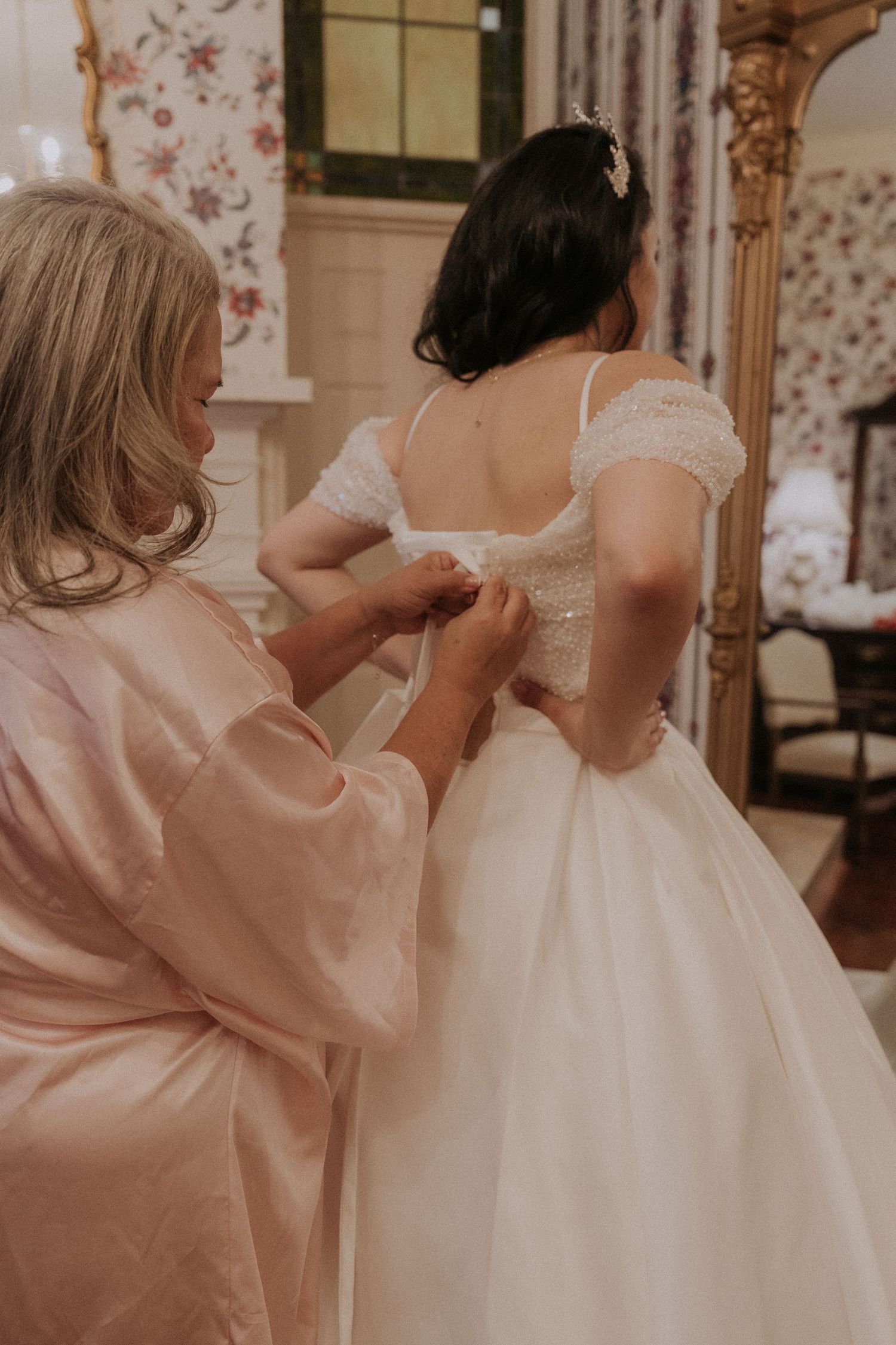 A woman in a pink robe fastens the back of a bride's white wedding gown in an elegant floral room.