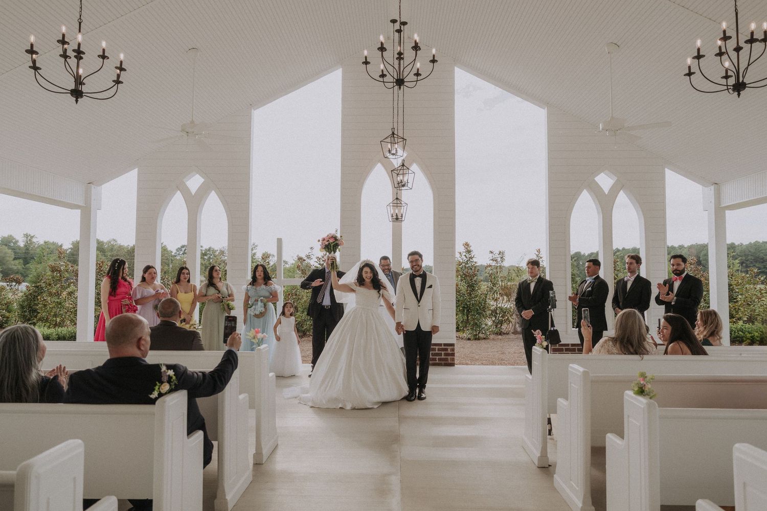 Bride and groom celebrate at altar surrounded by wedding party in stunning white chapel with arched windows.
