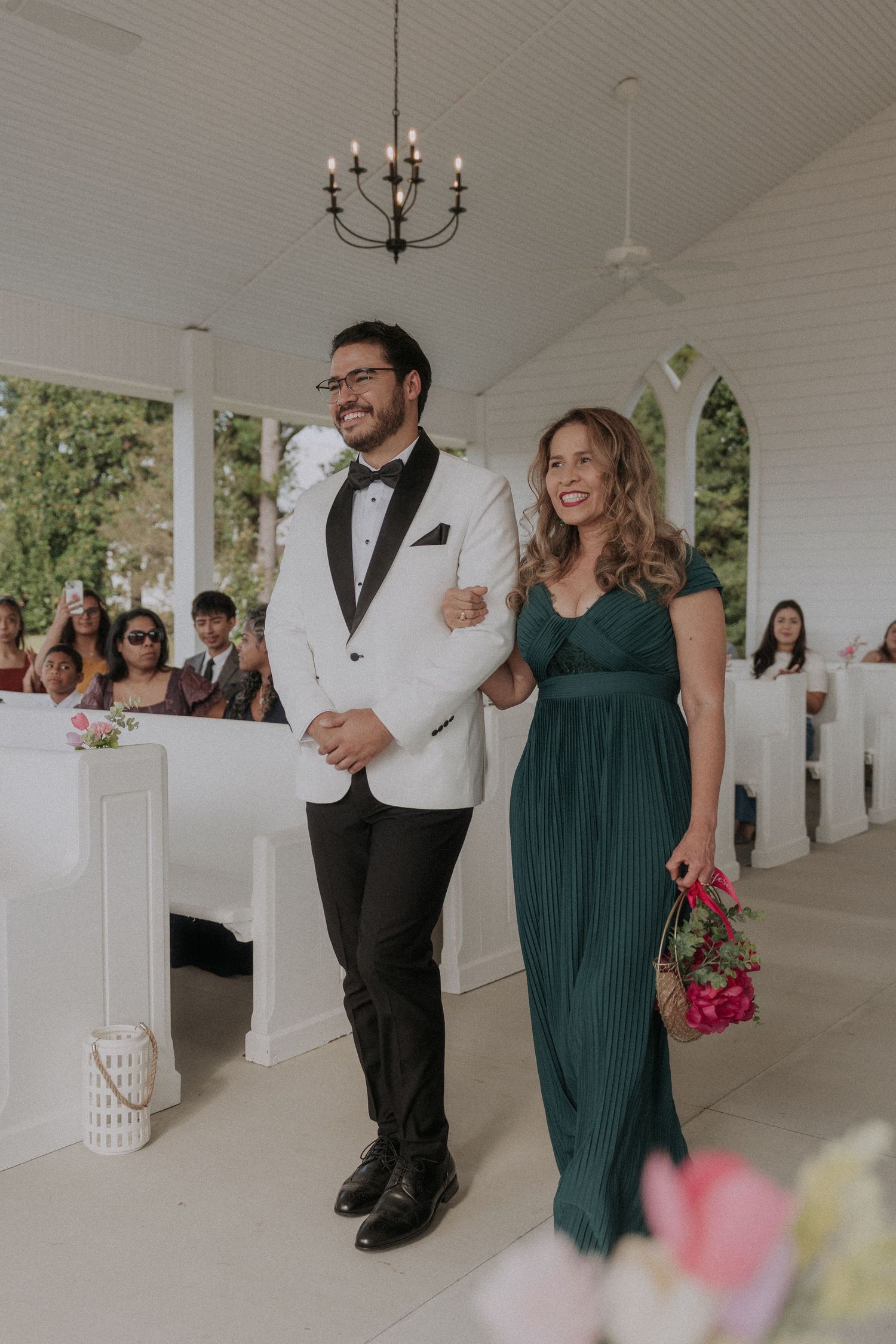 Groom in white tuxedo walks down aisle with bridesmaid in green gown at elegant white chapel wedding ceremony.