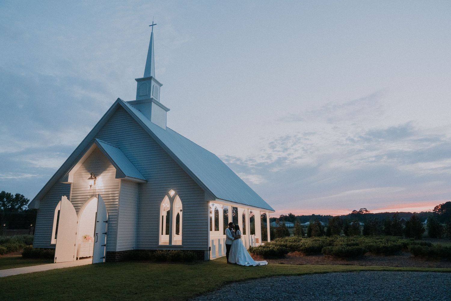 A bride stands outside a white chapel with a steeple at sunset, glowing warmly against a pastel evening sky.