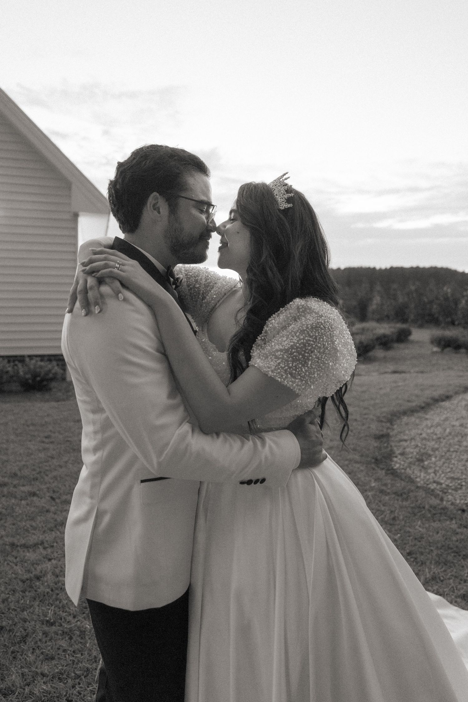 Black and white photo of a bride and groom embracing outdoors, nearly kissing, with a small building in the background.