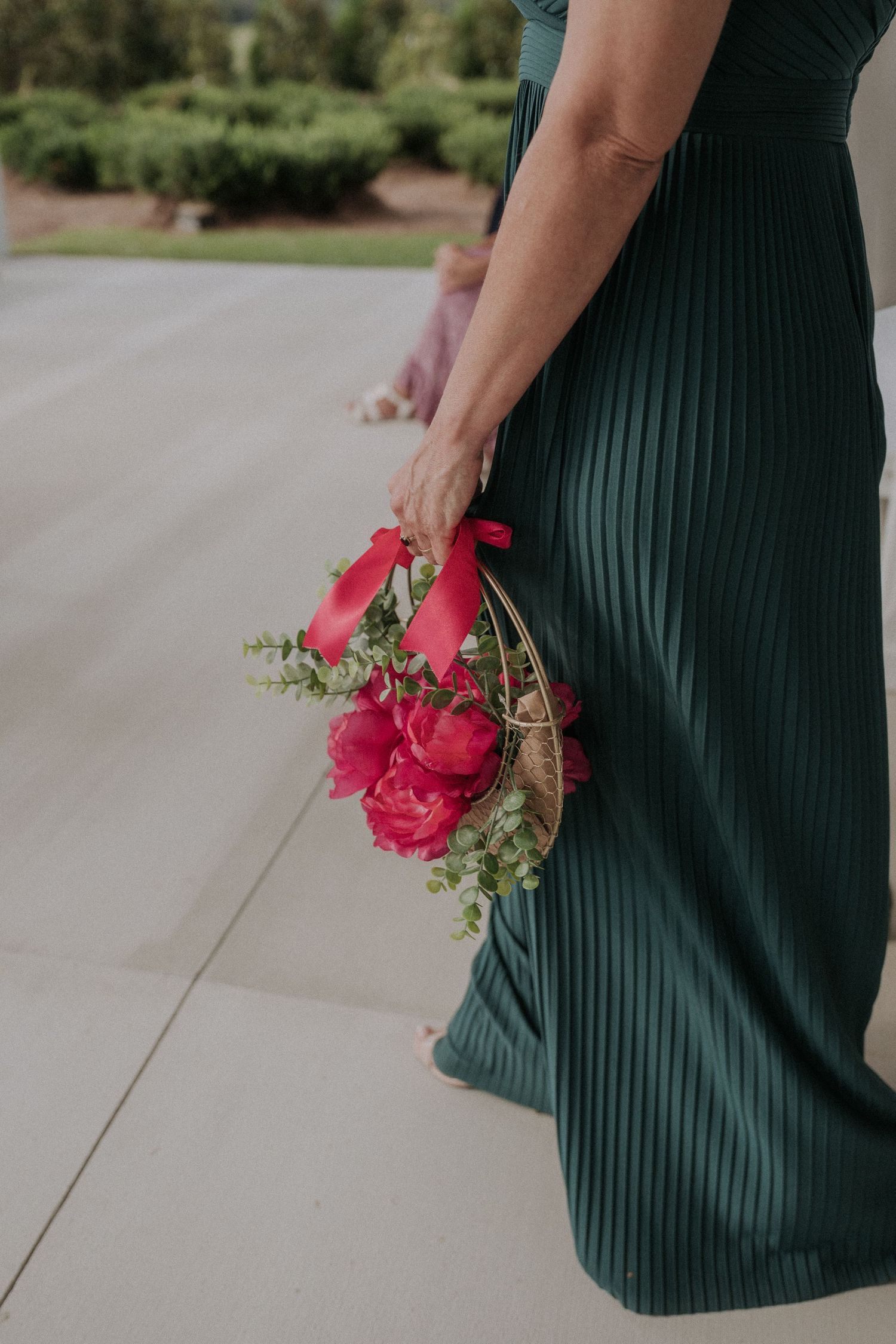 Bridesmaid in green pleated dress holding red peony bouquet with ribbon at outdoor wedding ceremony.