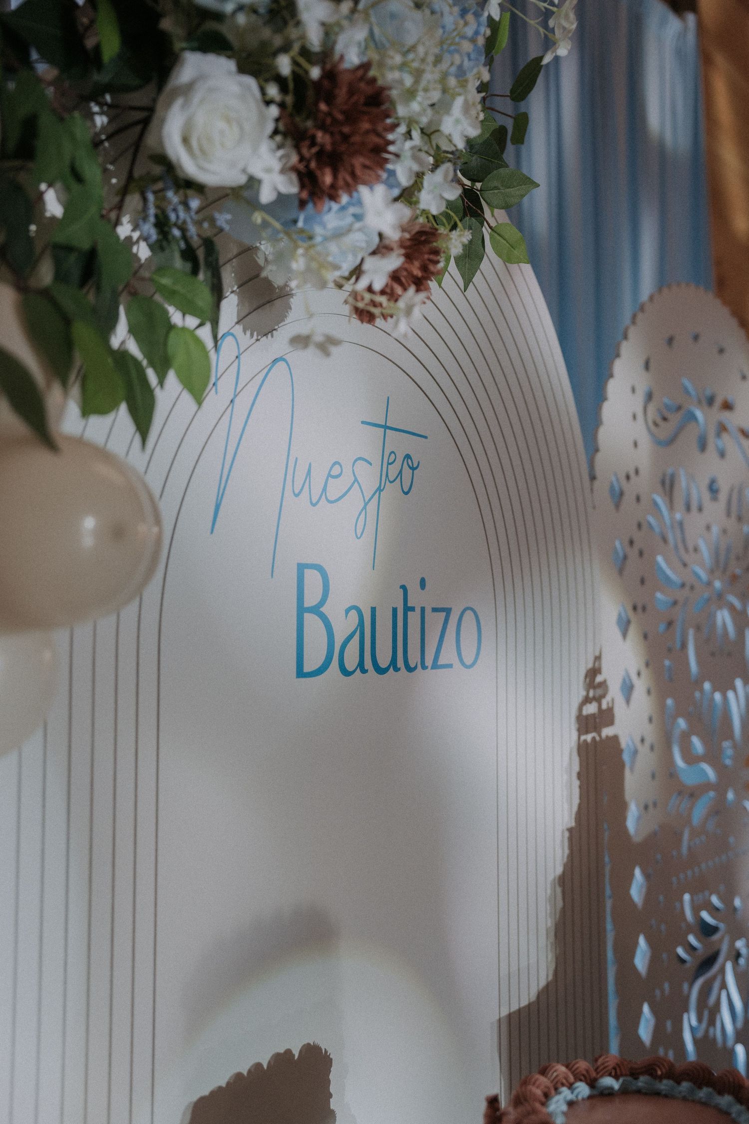 Elegant white decorative backdrop with floral arrangements and 'Bautizo' text in blue lettering.
