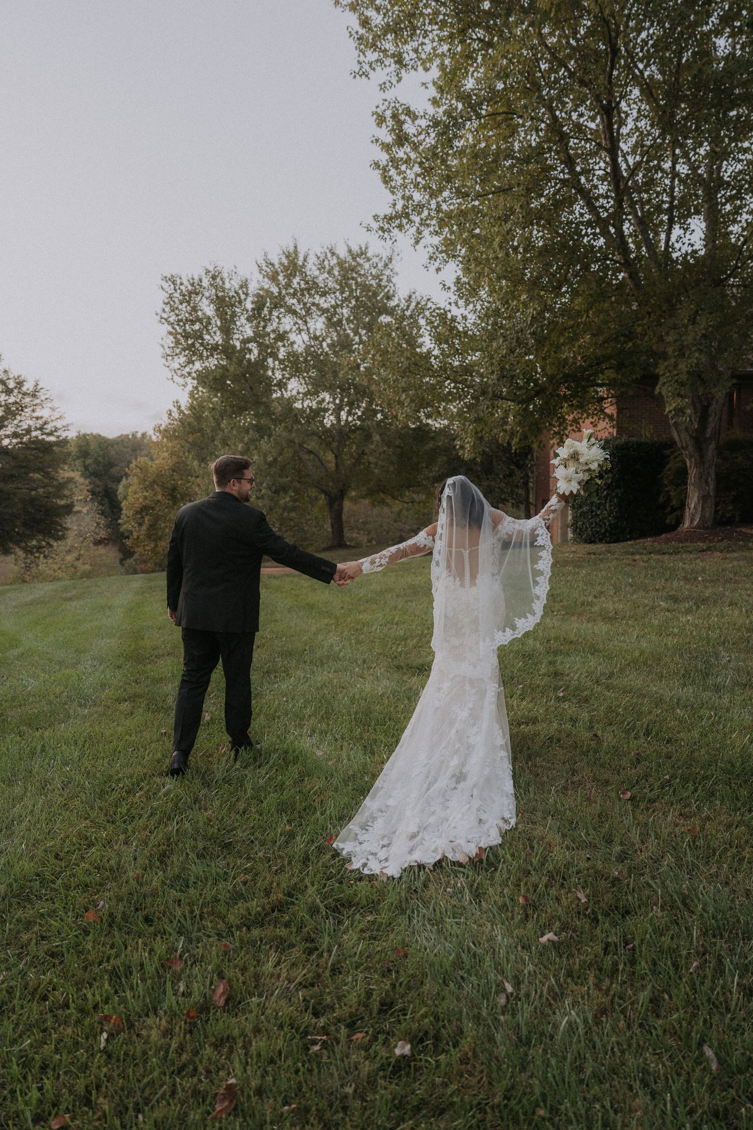 Couple in wedding attire playfully running through a grassy field at sunset.