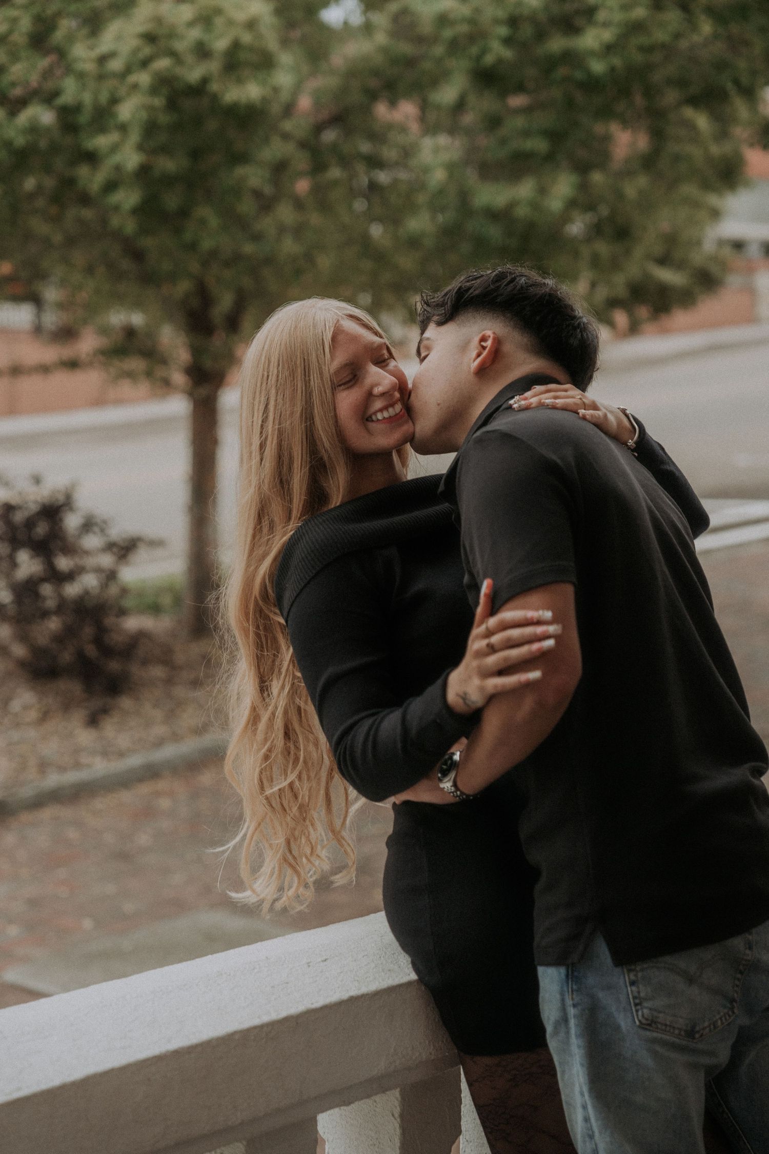 A couple in dark clothing share intimate moments together while leaning against an outdoor railing.