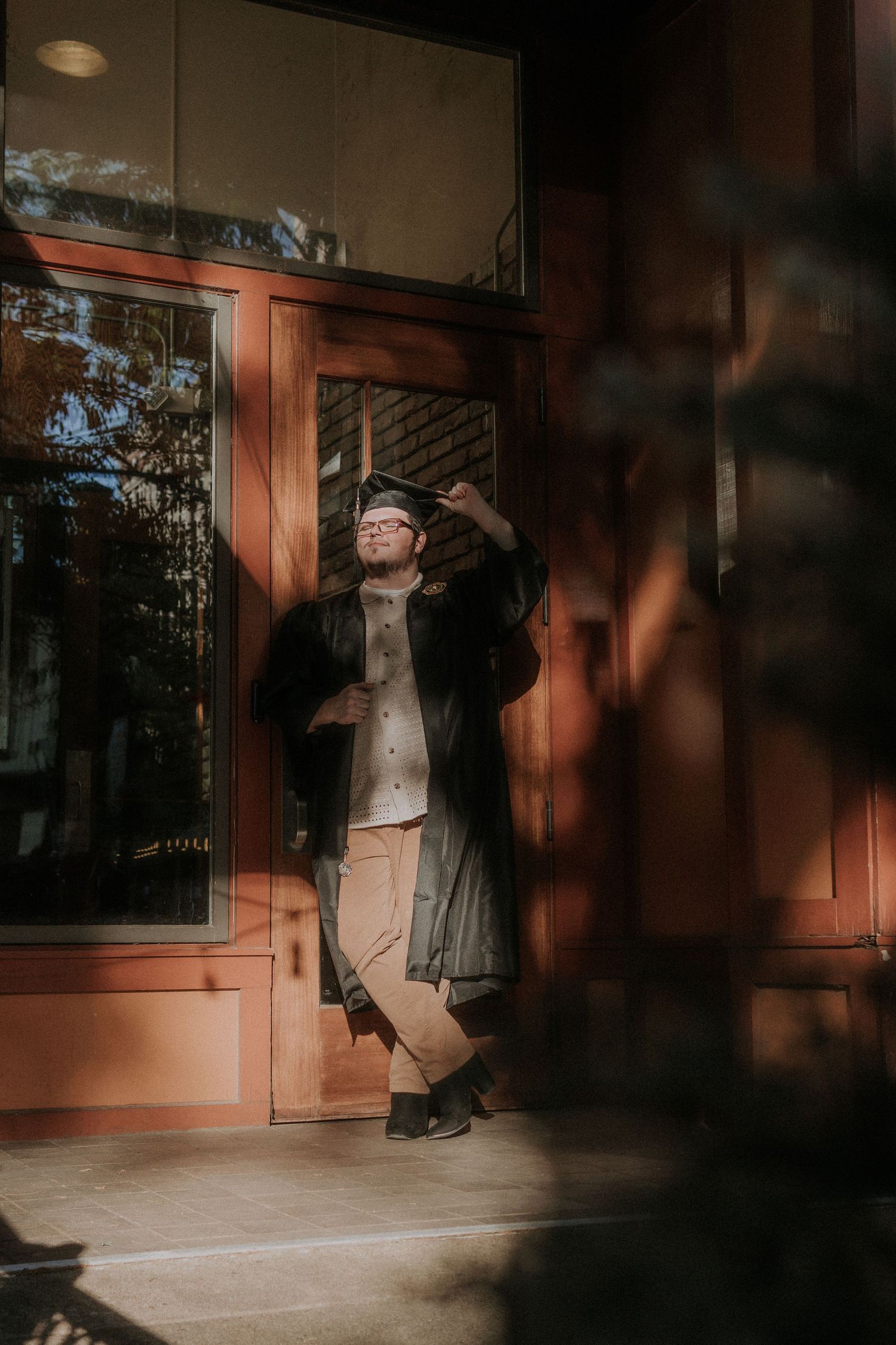 A figure in a graduation cap and gown poses dramatically against a wooden doorway in warm lighting.