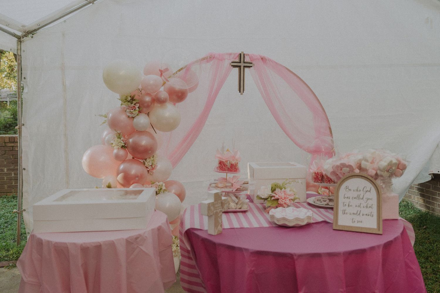 Pink and white party decorations with crosses and balloons set up under a tent.