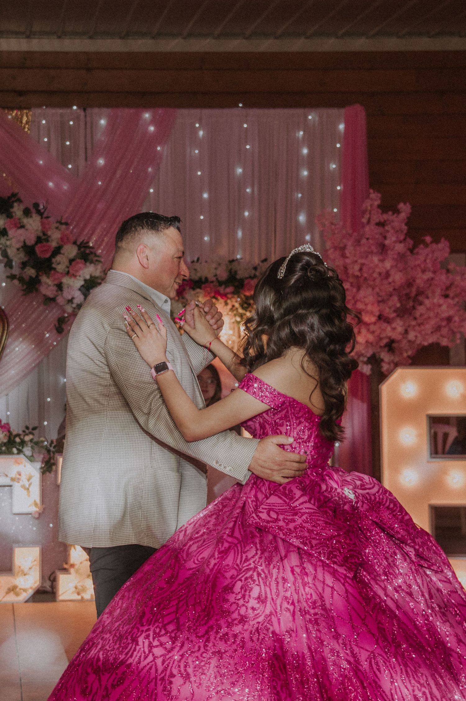 A godfather shares a dance at a Sweet 16 celebration with pink decor, flowers and twinkling lights in the background.