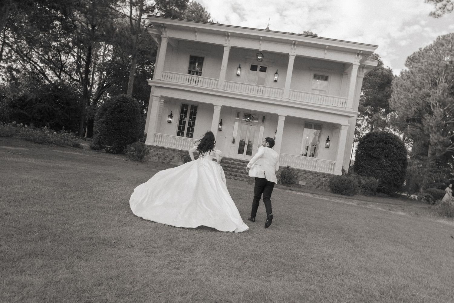 A couple in wedding attire runs across the lawn of an elegant white mansion.