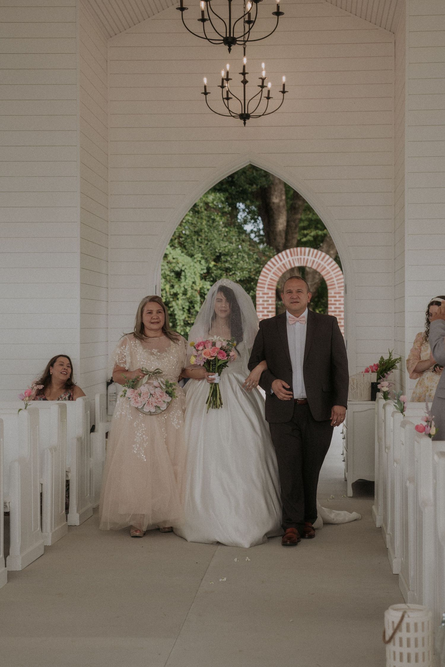 Bride walks down chapel aisle escorted by parents during traditional wedding ceremony.