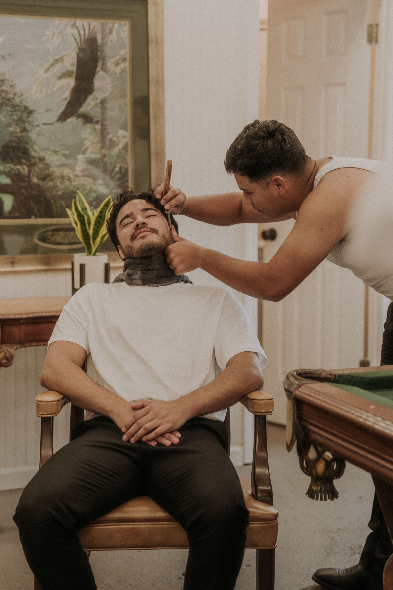 A barber carefully grooms a seated man wearing a neck brace in a vintage-styled room with a pool table.