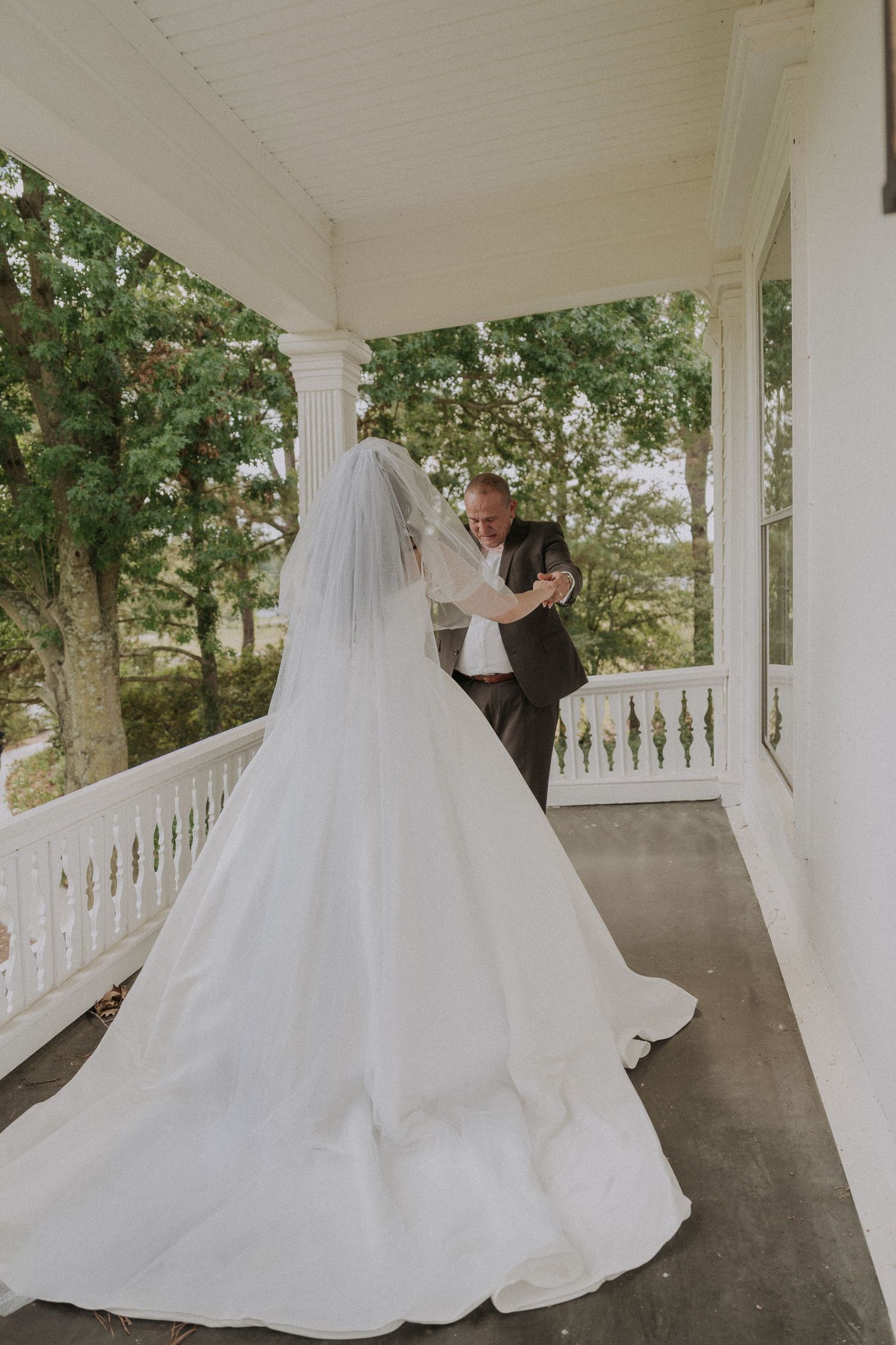 Bride in white gown with veil shares first look moment with father on elegant porch.