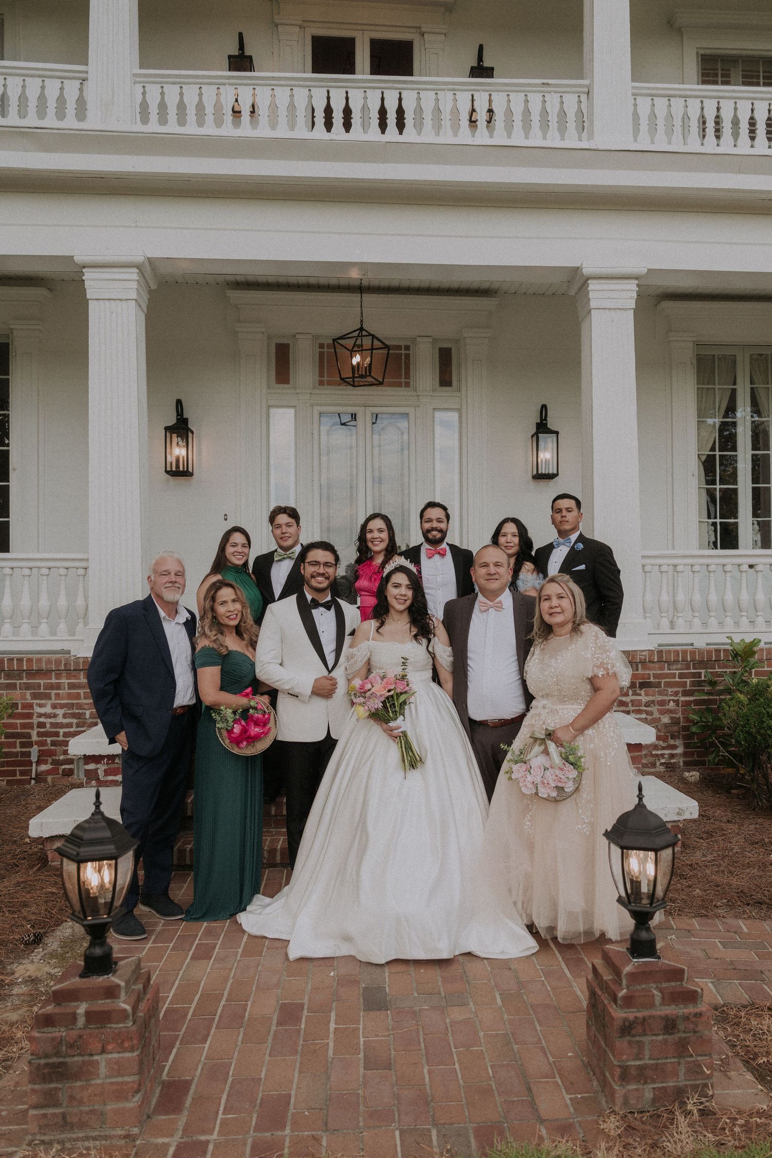 Wedding party poses in front of elegant white plantation-style venue with brick steps and balcony.