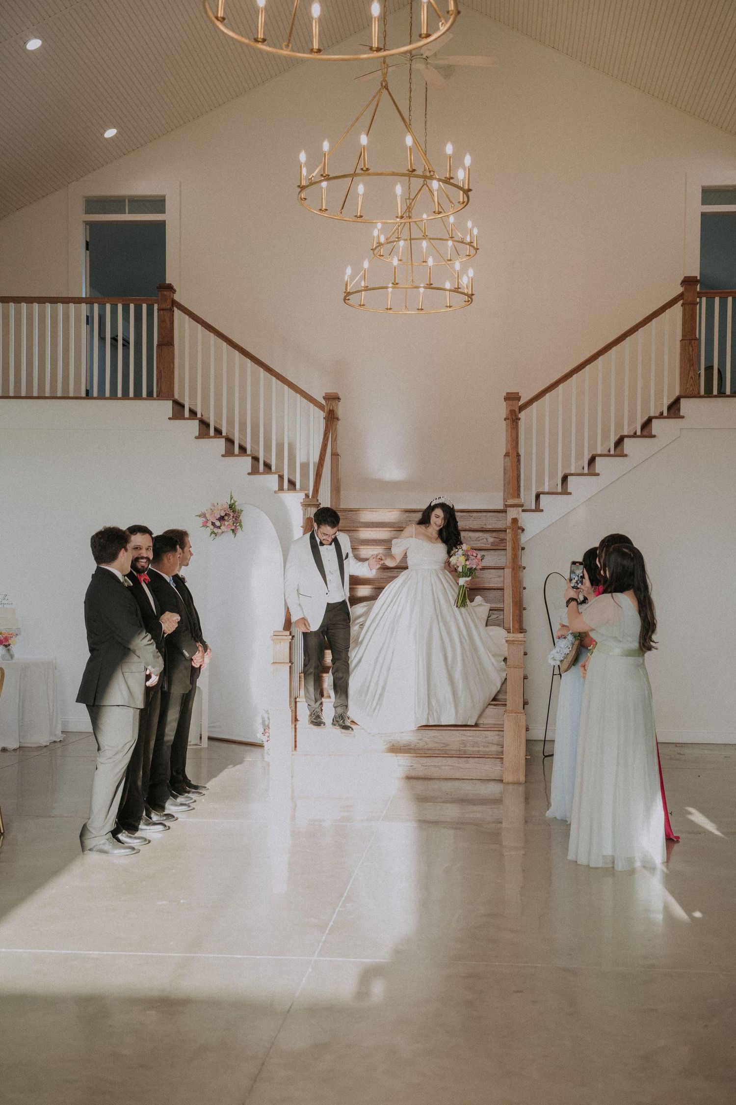 Bride and groom stand between dual staircases under gold chandeliers in elegant white wedding venue.