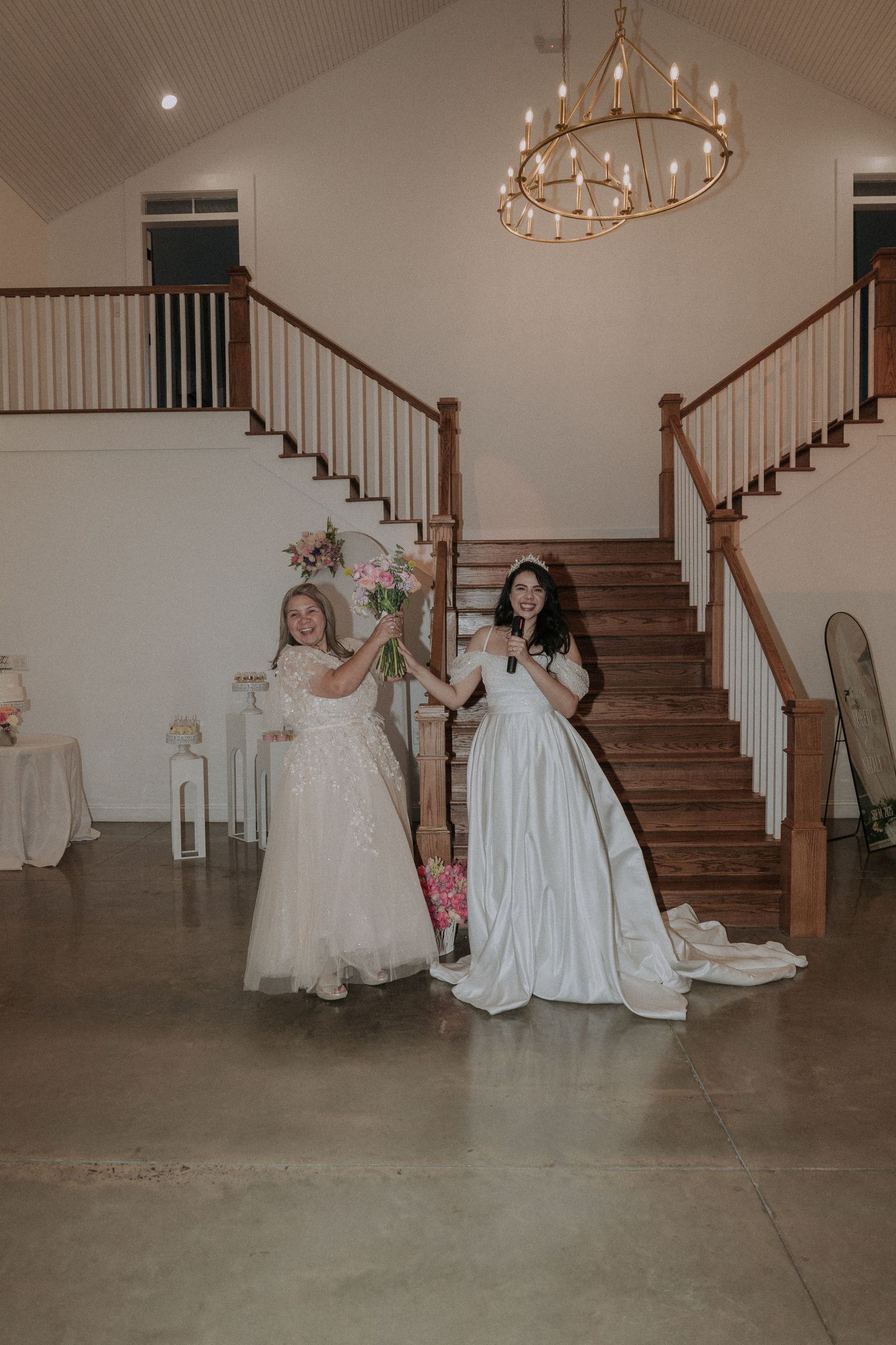 Bride in white gown poses with flower girl near grand staircase in elegant wedding venue with chandelier.