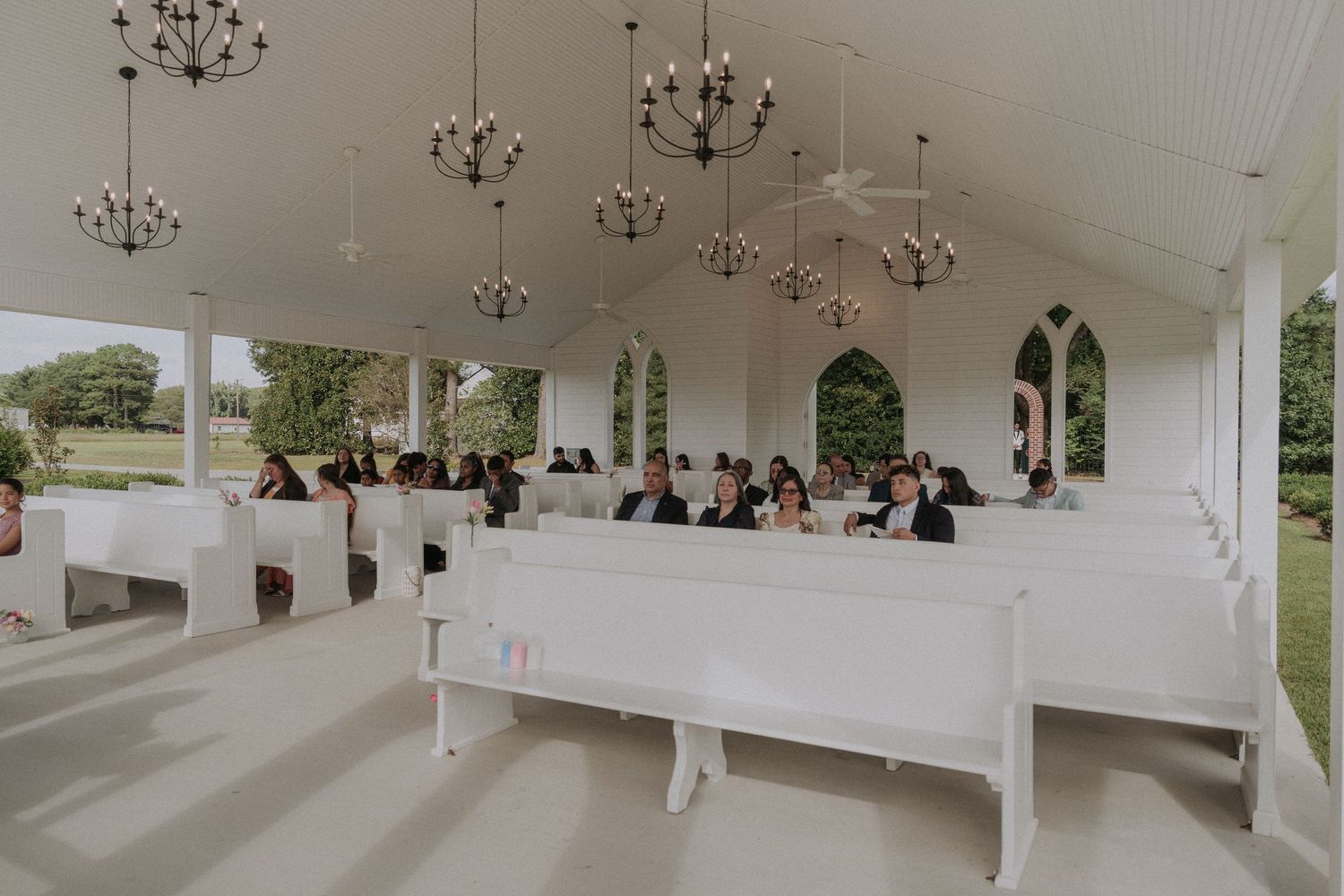Elegant open-air wedding chapel with white pews, chandelier lighting, and guests seated for a ceremony.