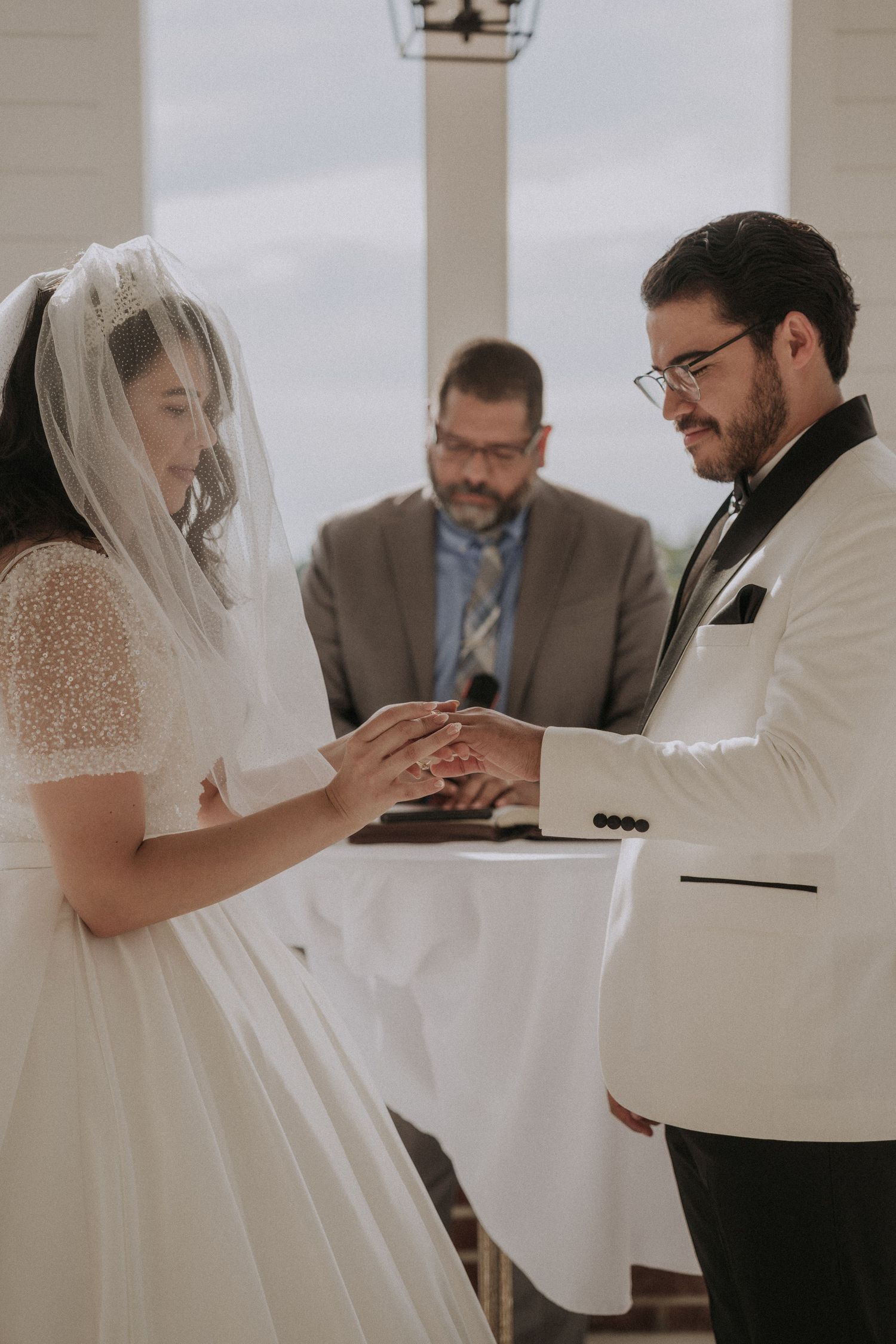 Groom placing wedding ring on brides finger during ceremony with officiant in background.