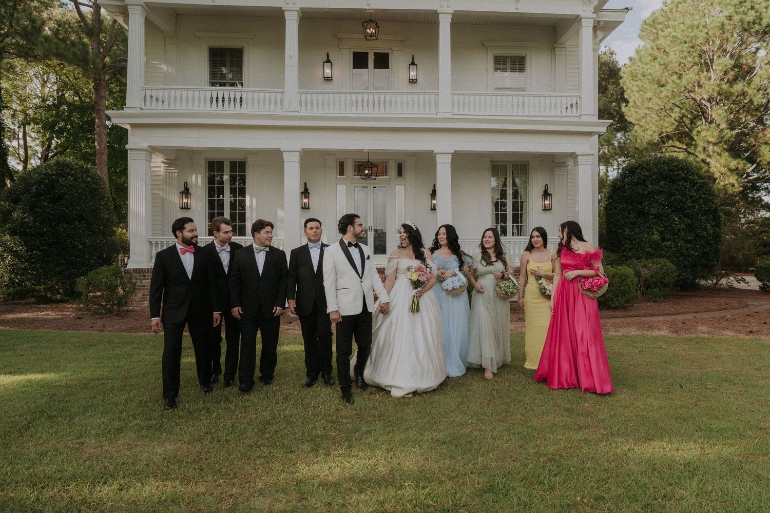 Wedding party with bride, groom, groomsmen in black suits, and bridesmaids in colorful gowns pose on lawn of white estate.