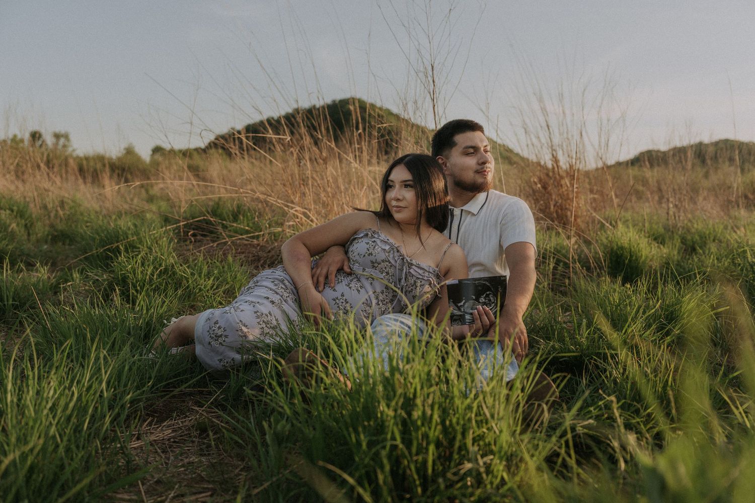 Couple sitting together in tall green grass field at golden hour, woman in floral dress leaning on man in white polo shirt.