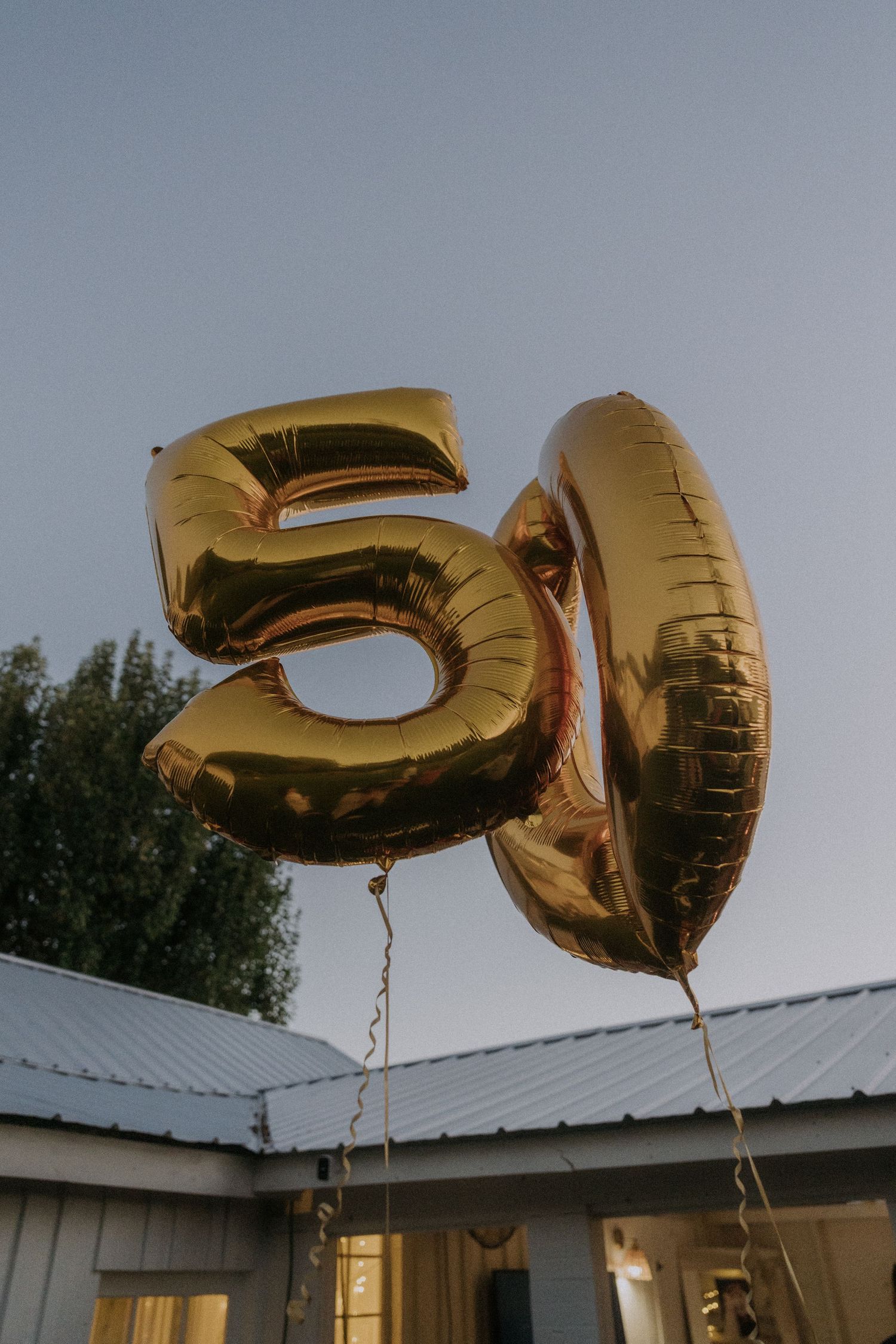 Gold number 50 balloons floating against an evening sky with house in background.