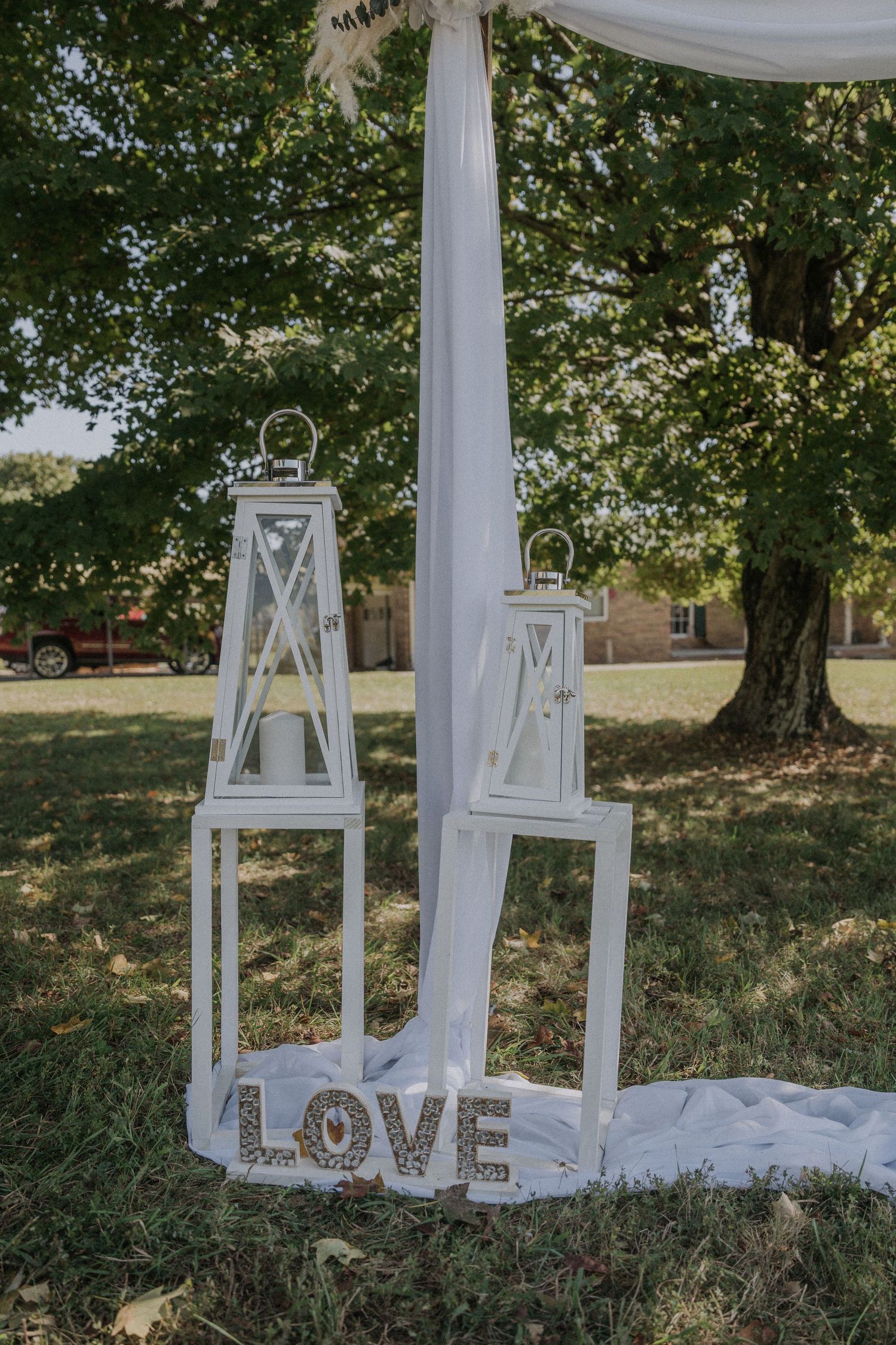 Romantic outdoor wedding setup with white lanterns and 'LOVE' sign under draped fabric and trees.