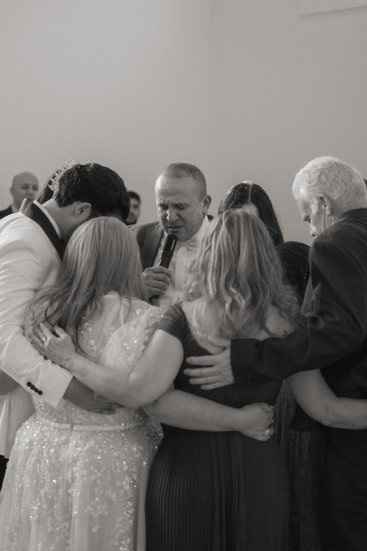 Black and white photo of wedding party gathered in group prayer with officiant holding microphone.