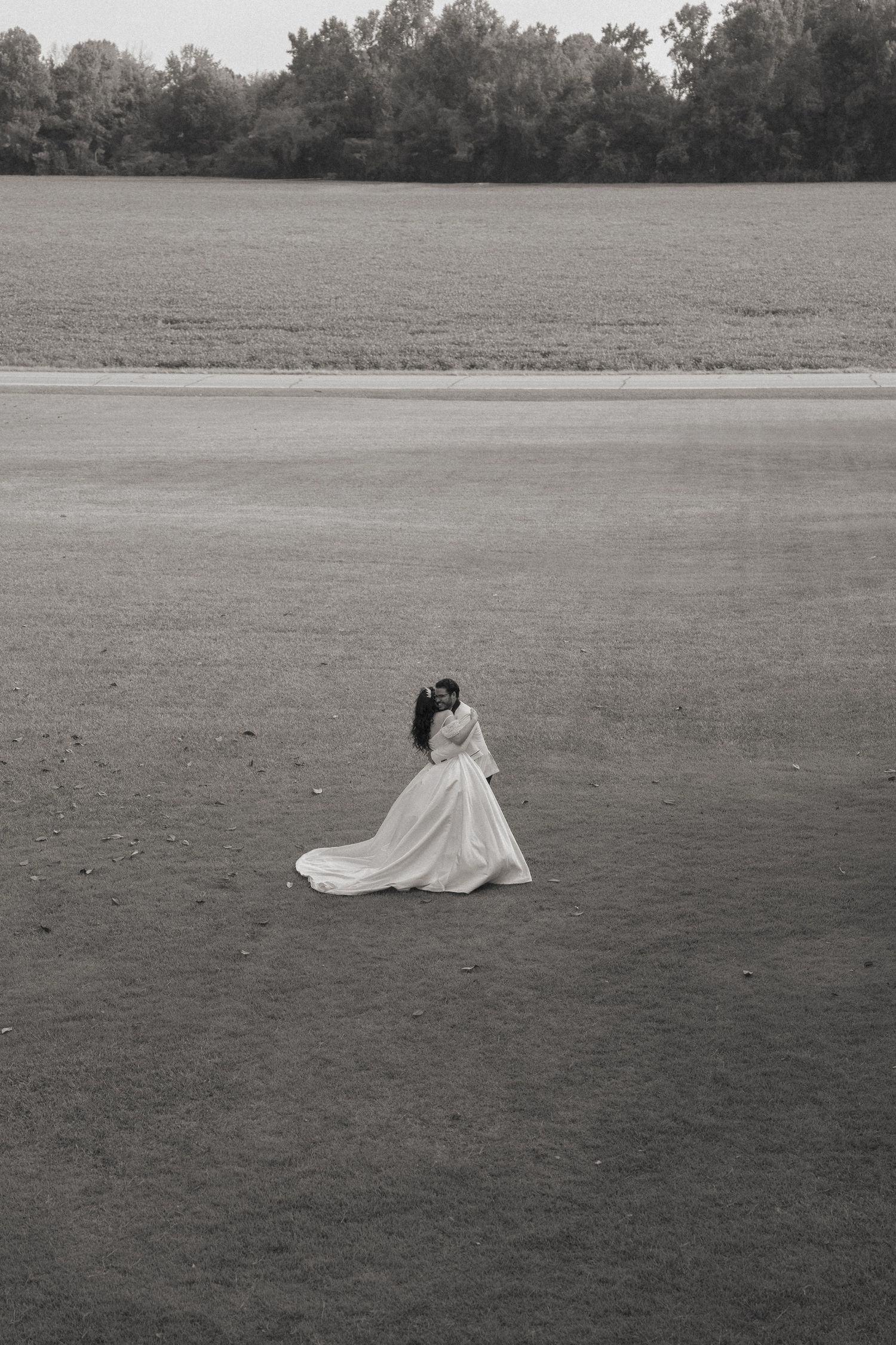 Black and white photo of bride and groom embracing in an open field with trees in the background.