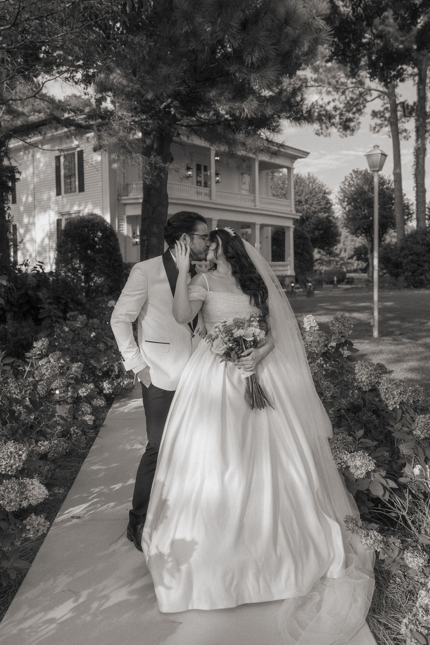 Black and white photo of a bride and groom sharing a romantic moment on a garden path outside an elegant estate.