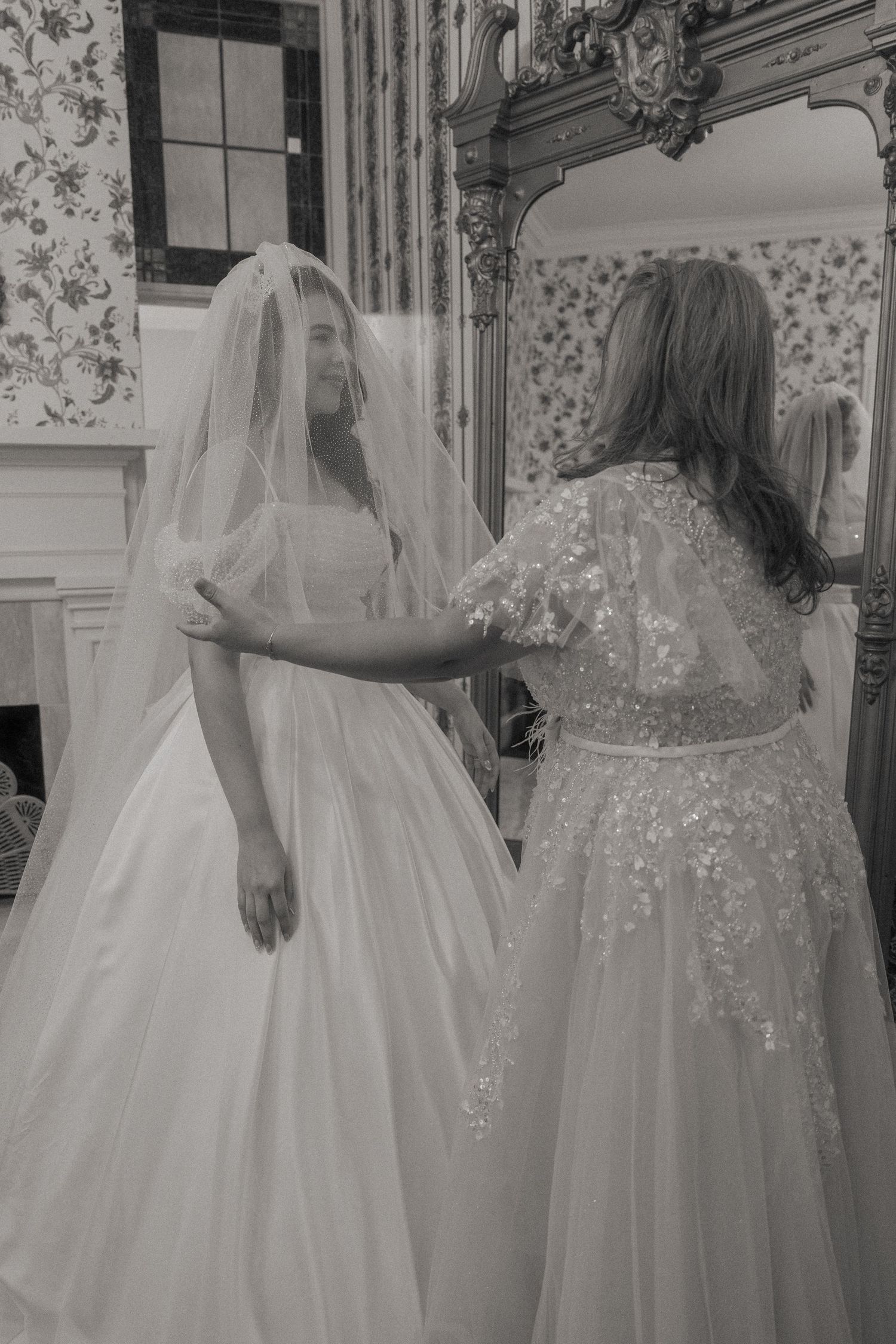 Bride in white gown and veil assisted by woman in lace dress near ornate mirror, black and white photo.