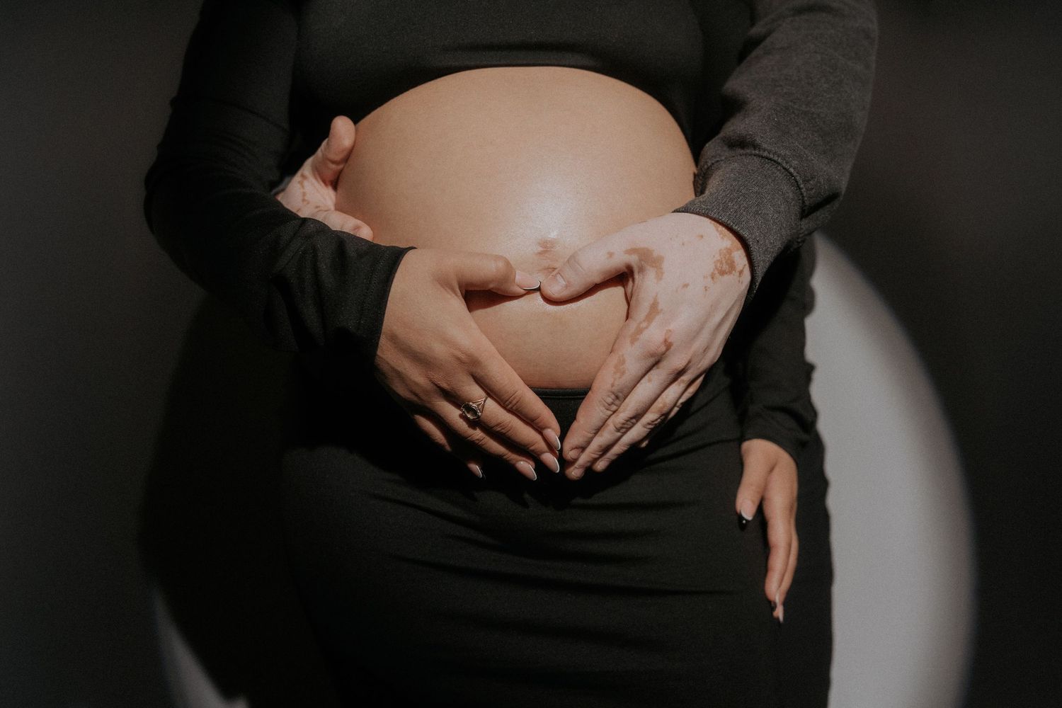 Maternity photoshoot showing close up shots of a pregnant belly in black dress against dark background.
