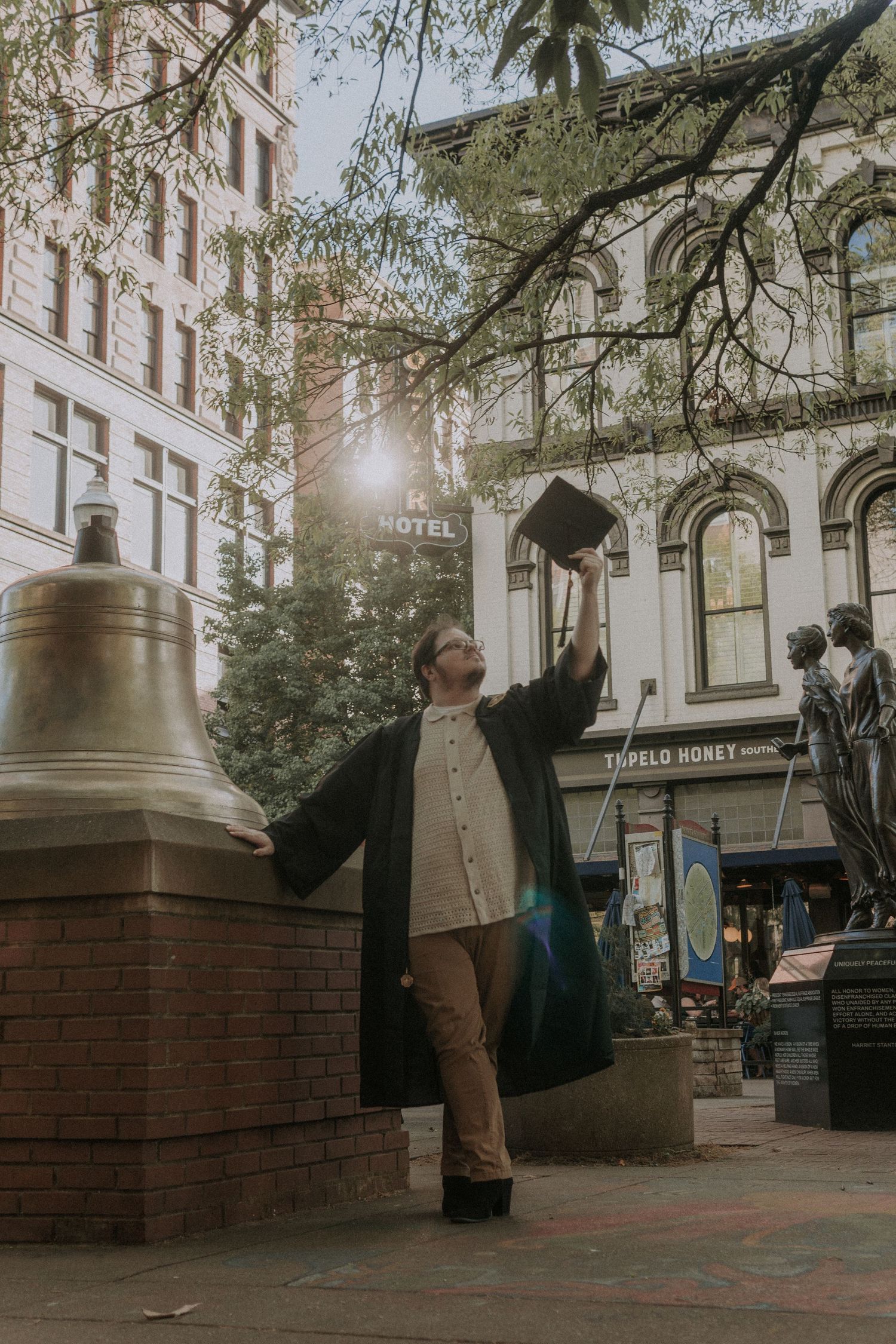 A male graduate stands next to a large bell in front of a historic hotel building on a city street.