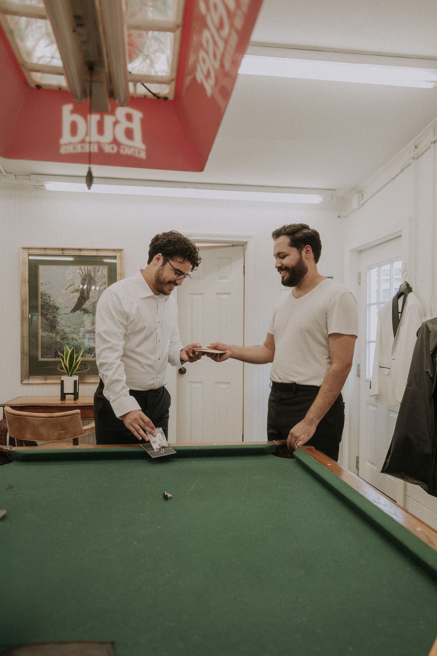 Two men smiling and exchanging cards by a pool table under a Budweiser sign.