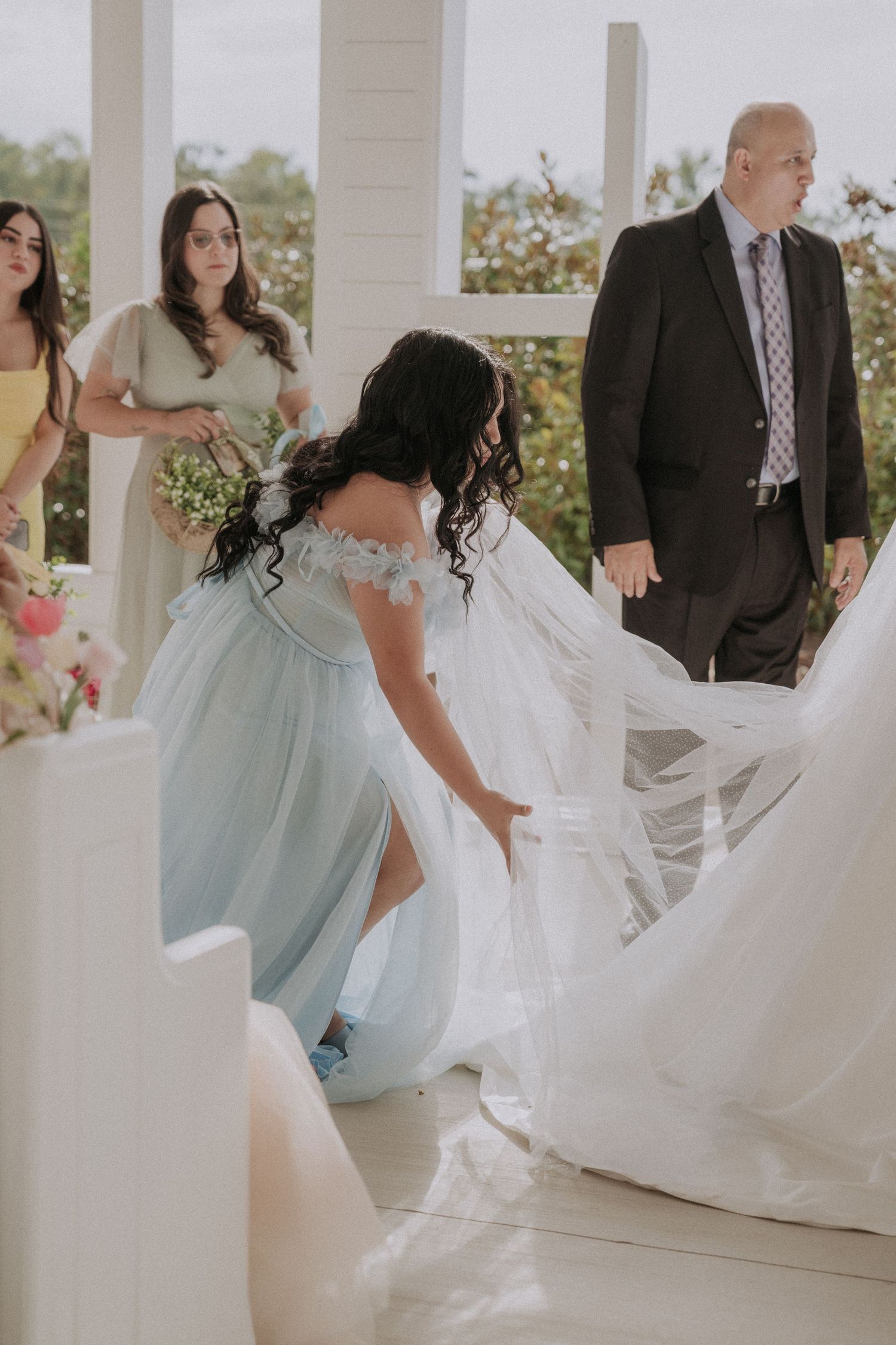 Bridesmaid in light blue gown adjusting bride's veil at outdoor wedding ceremony with guests in background.