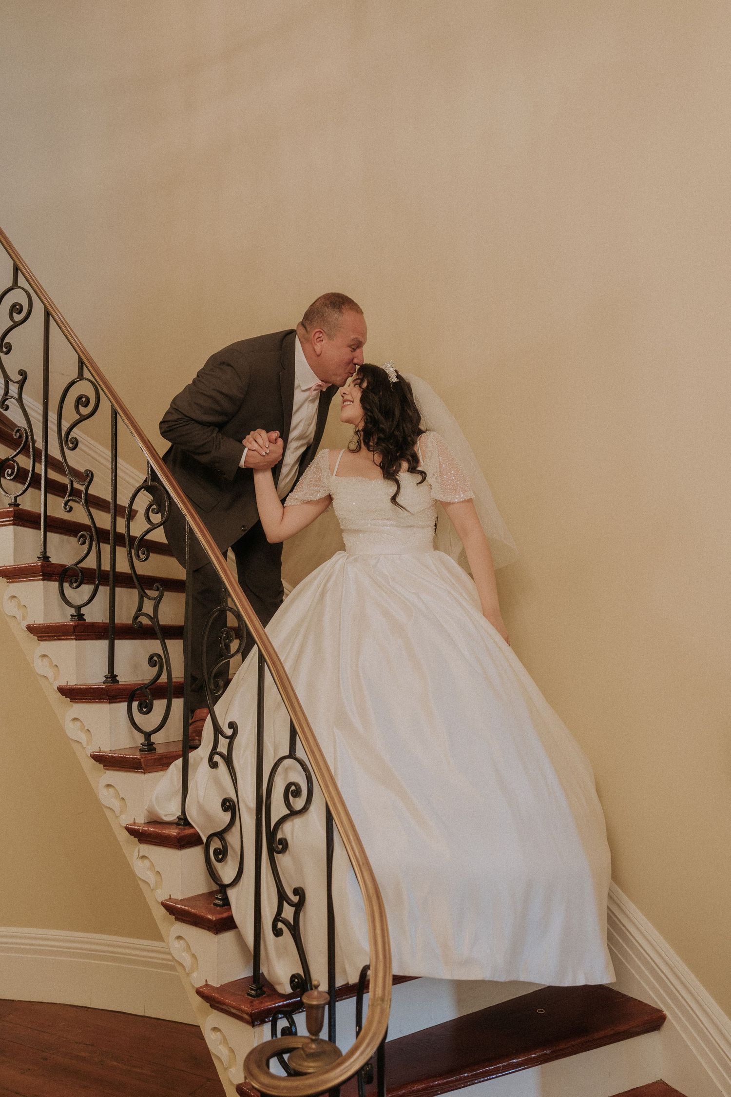 Bride and groom share a romantic kiss on an elegant curved staircase with wrought iron railing on their wedding day.