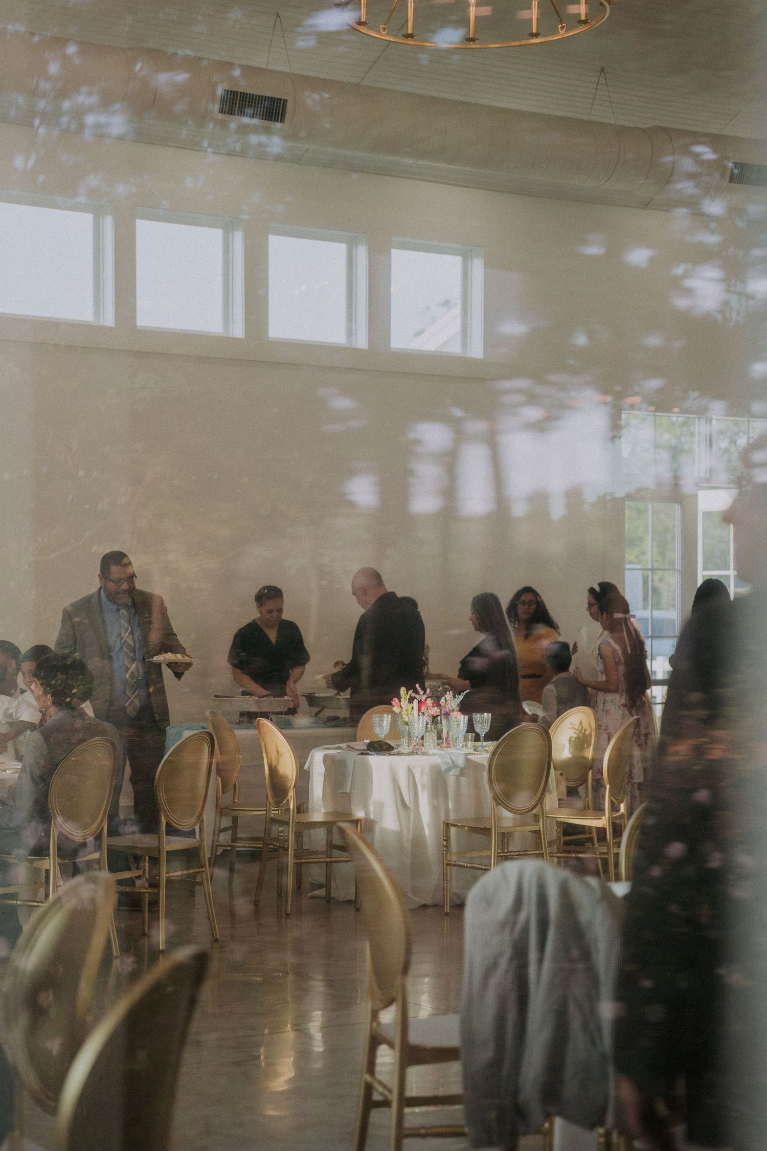 Elegant wedding reception venue with guests mingling around white-clothed tables and gold chairs in a bright, airy room.